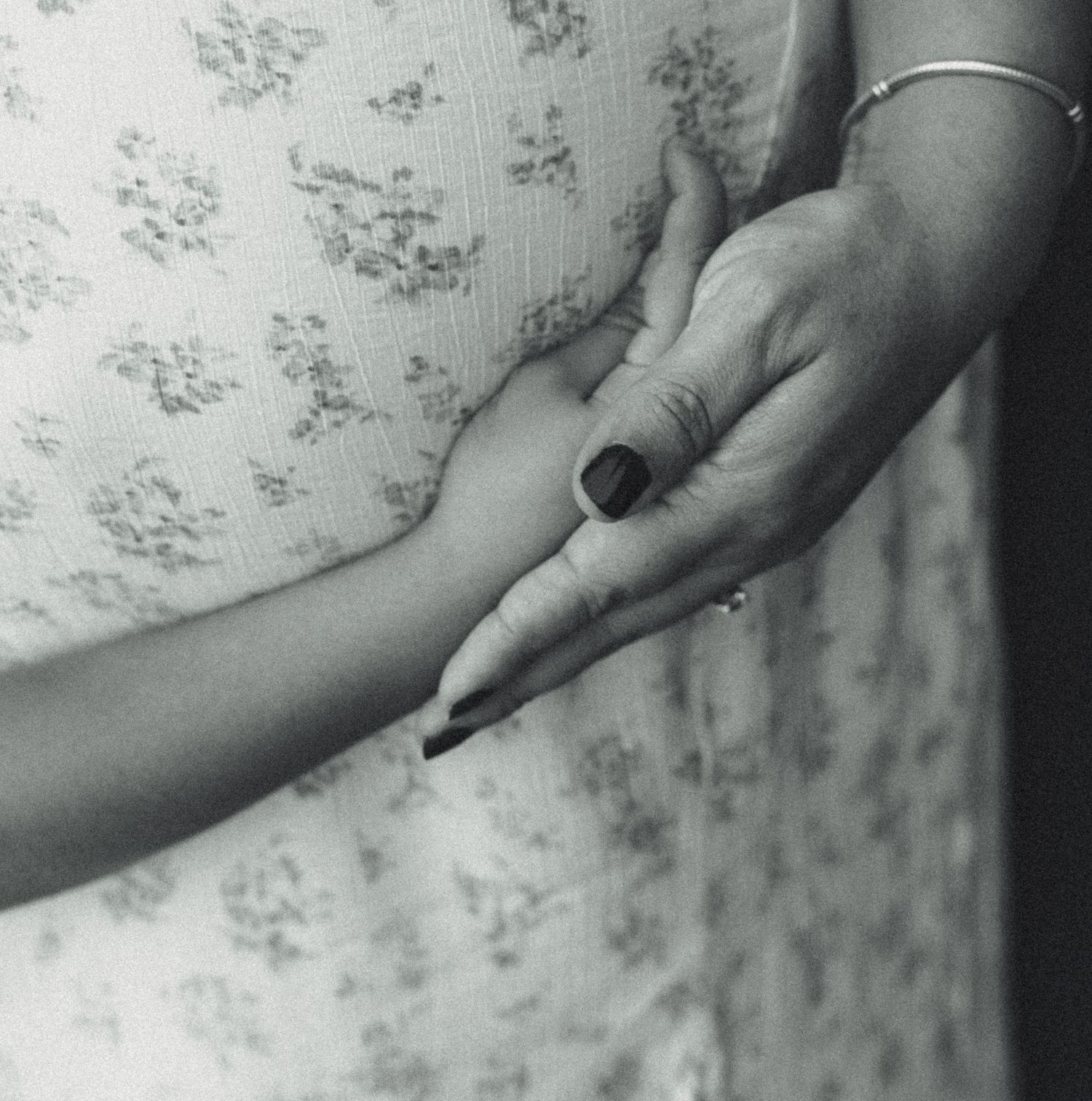 Black and white photo of two hands holding, one with dark nail polish and a bracelet, against a floral fabric background.