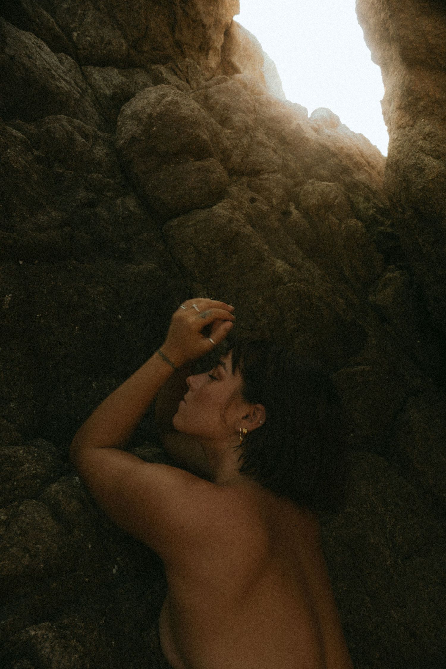 Woman resting against rocky cave walls with dramatic light streaming through an opening above.