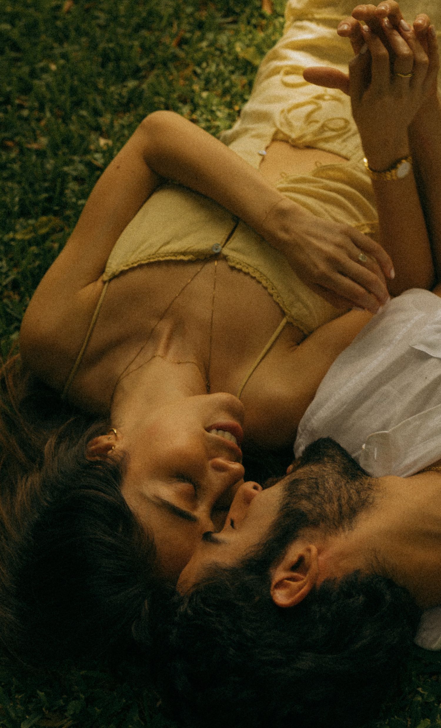 A couple lies on green grass, sharing an intimate moment, the woman in a yellow dress and the man in white.