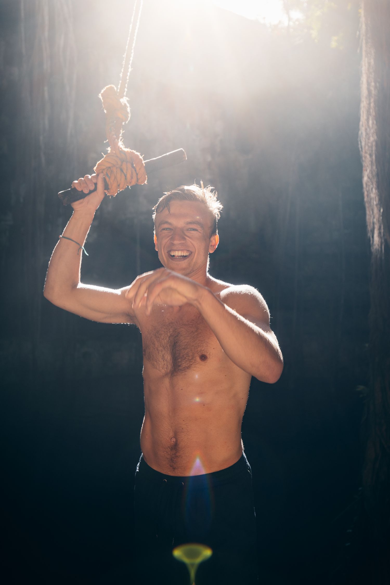 Shirtless muscular man smiling while holding a rope in sunlit forest.