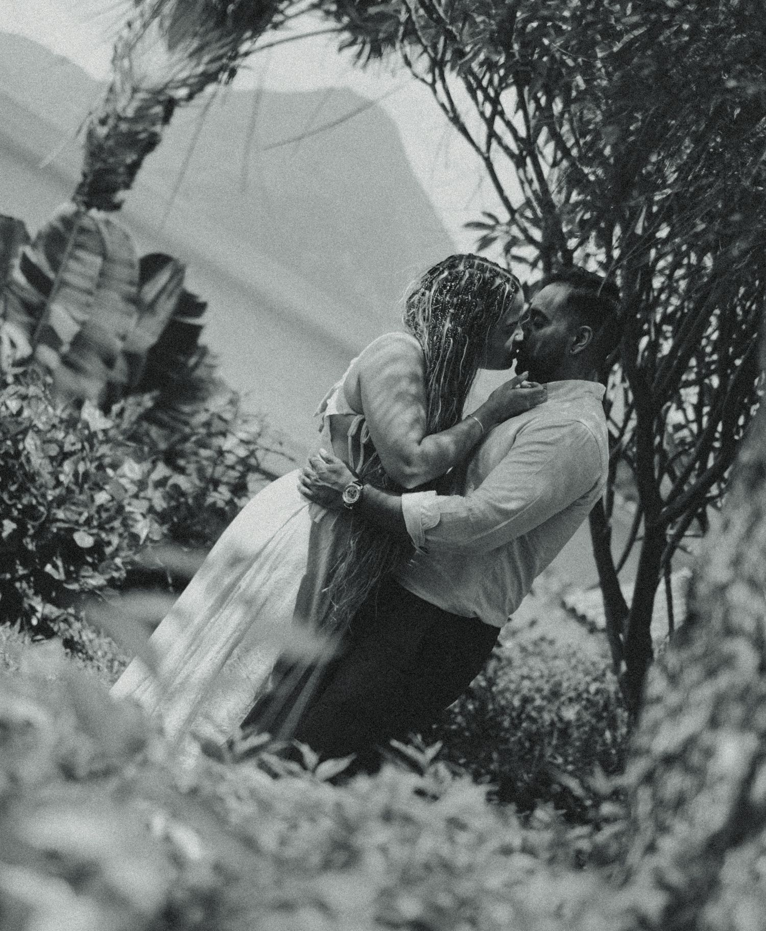 Black and white photo of a couple sharing a romantic kiss surrounded by lush tropical greenery and trees.