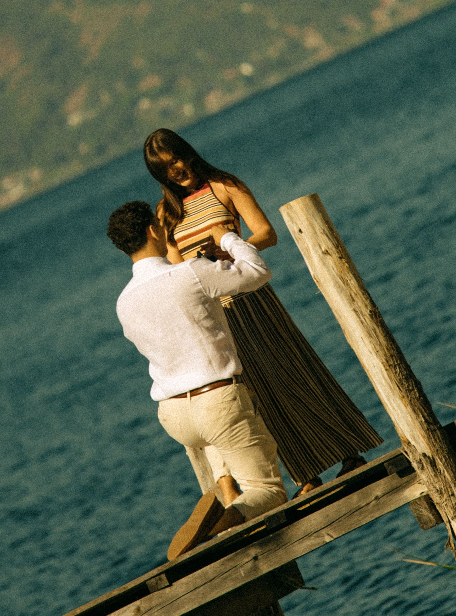 Couple embracing on a wooden dock by the water at golden hour, woman in striped top and pleated skirt.