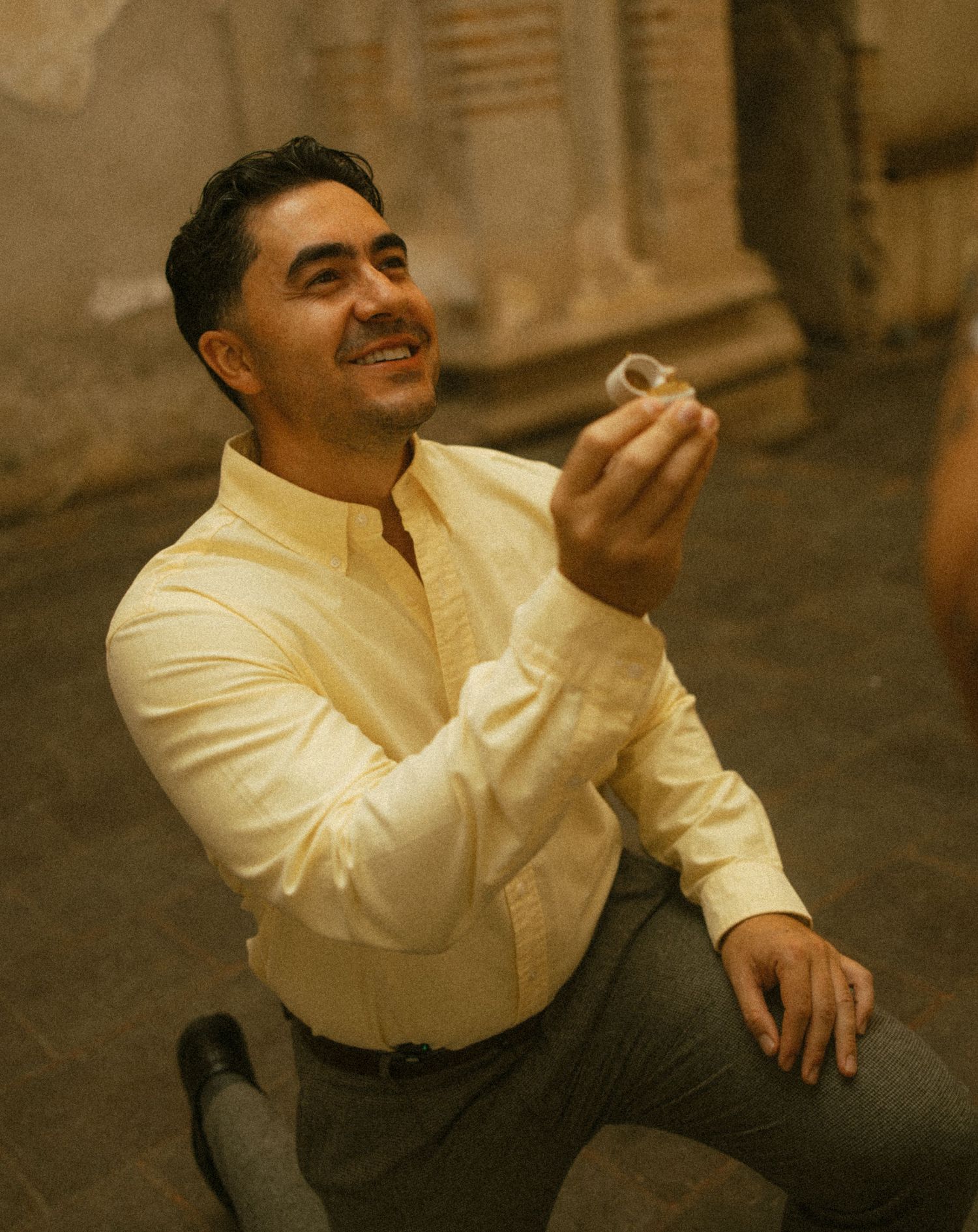 Smiling man in a yellow shirt kneels on one knee holding up a ring in a romantic proposal inside a rustic stone building.