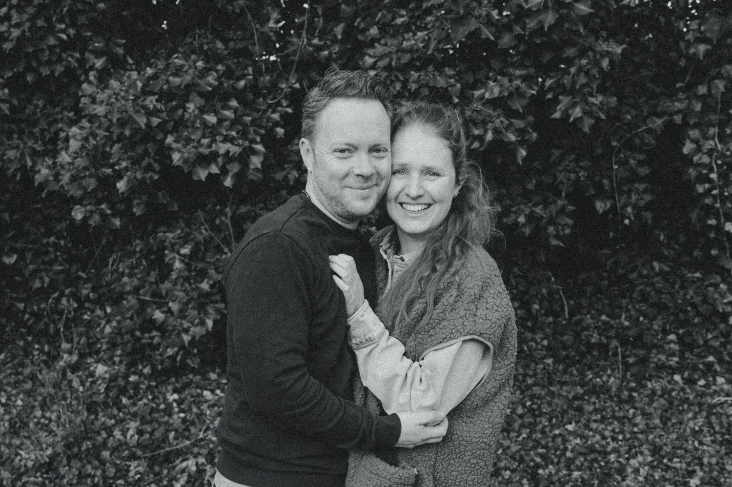 Black and white photo of a smiling couple embracing outdoors in front of a lush ivy-covered wall.