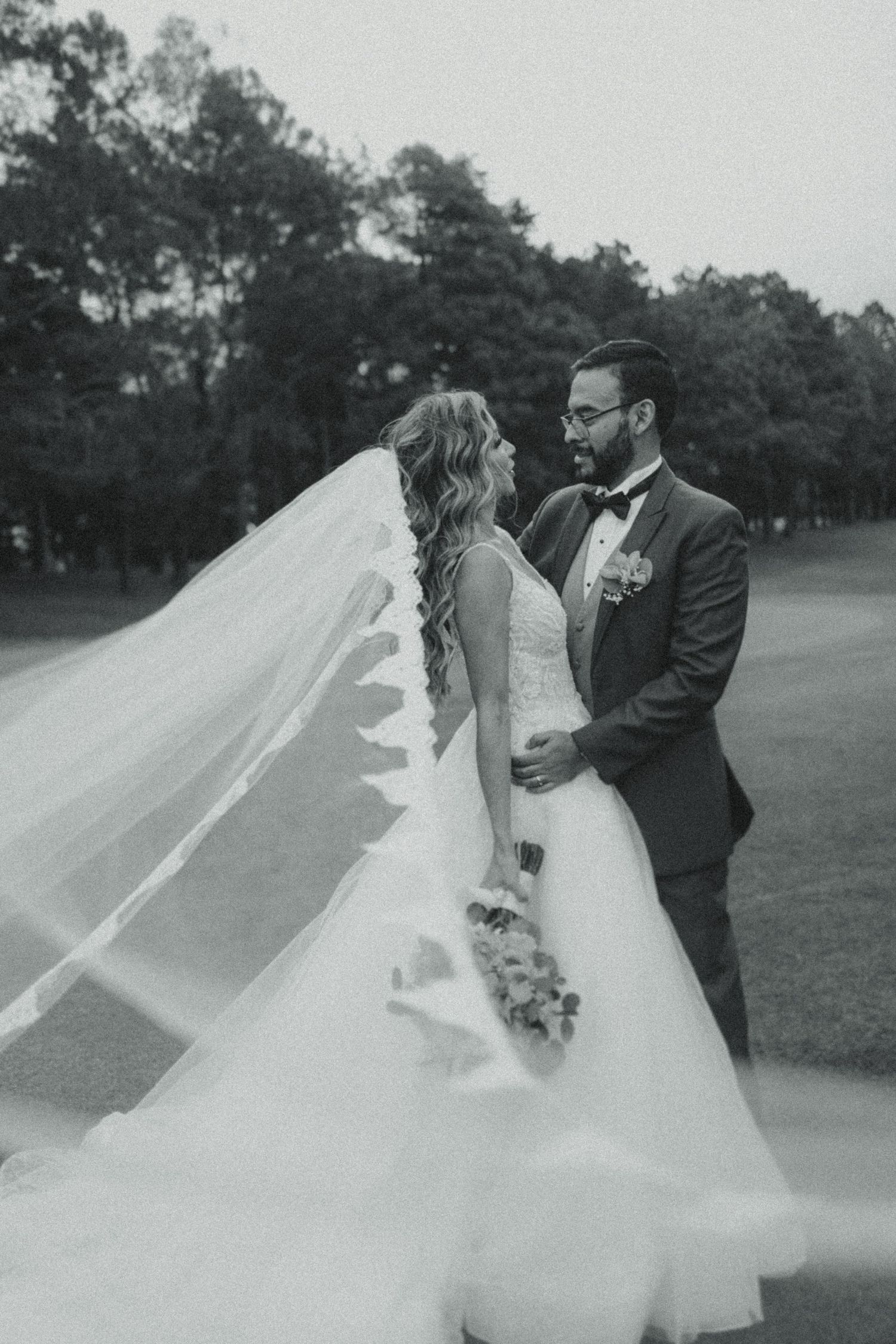 Black and white photo of a bride and groom sharing a romantic moment, her veil flowing gracefully on a golf course.