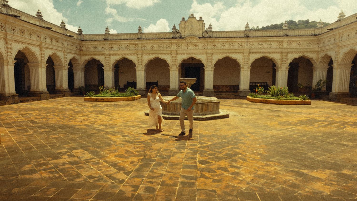 Couple dancing in a historic colonial courtyard with arched corridors and a central fountain under a blue sky.