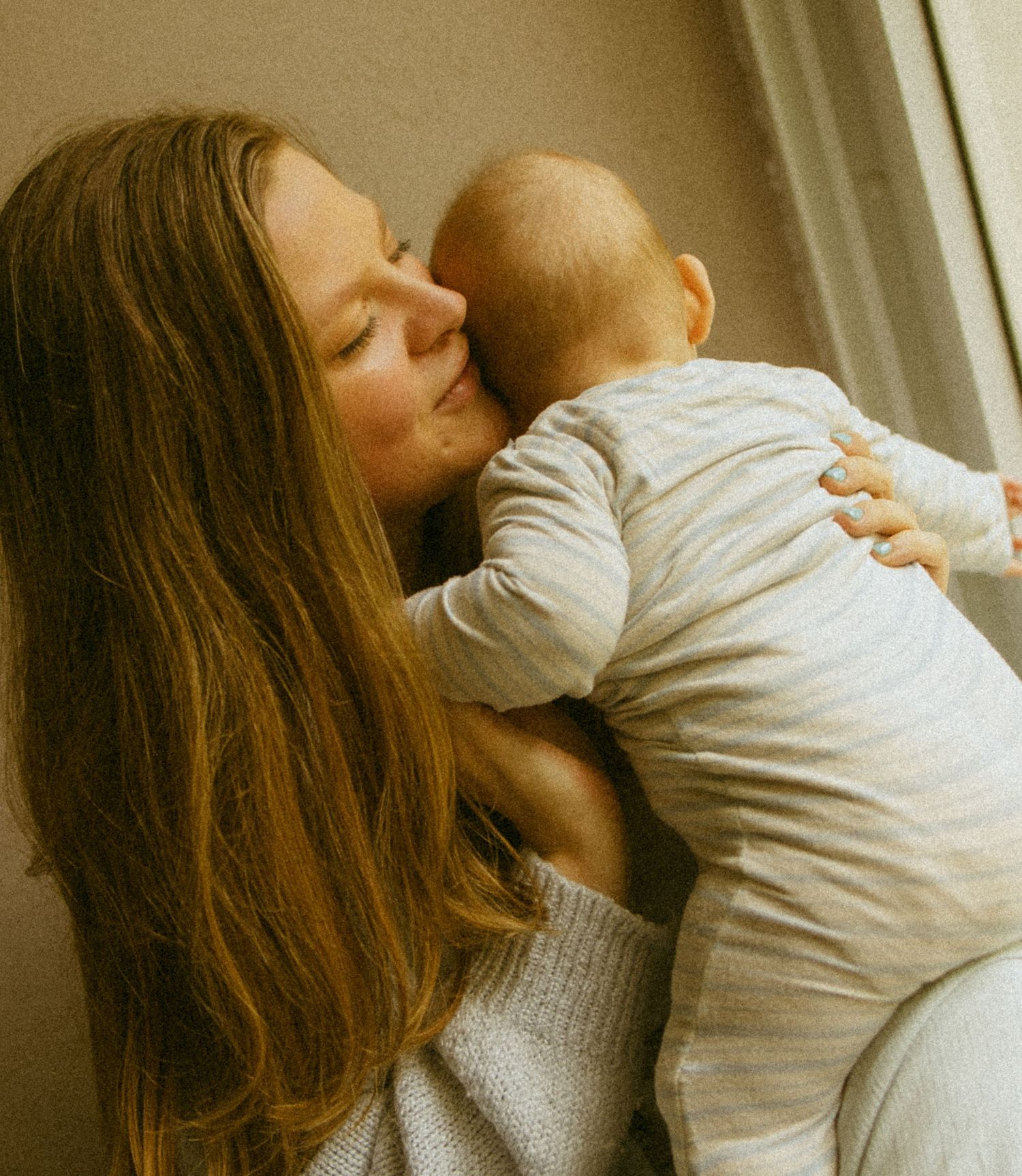 A tender moment as a young mother with long hair gently kisses her infant baby dressed in a soft grey onesie.