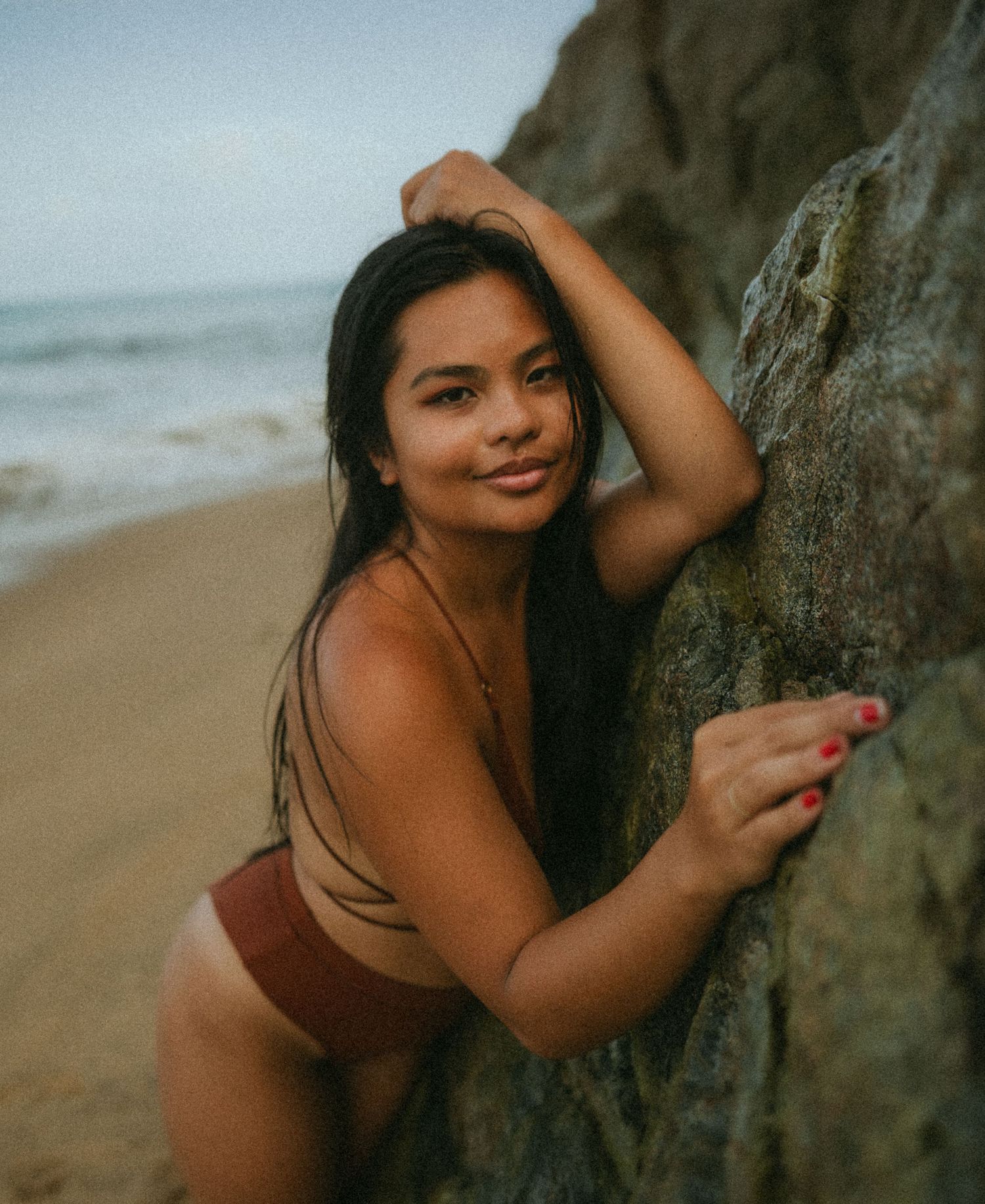 Young woman in a brown bikini posing against a rocky cliff on a sandy beach with ocean waves in the background.