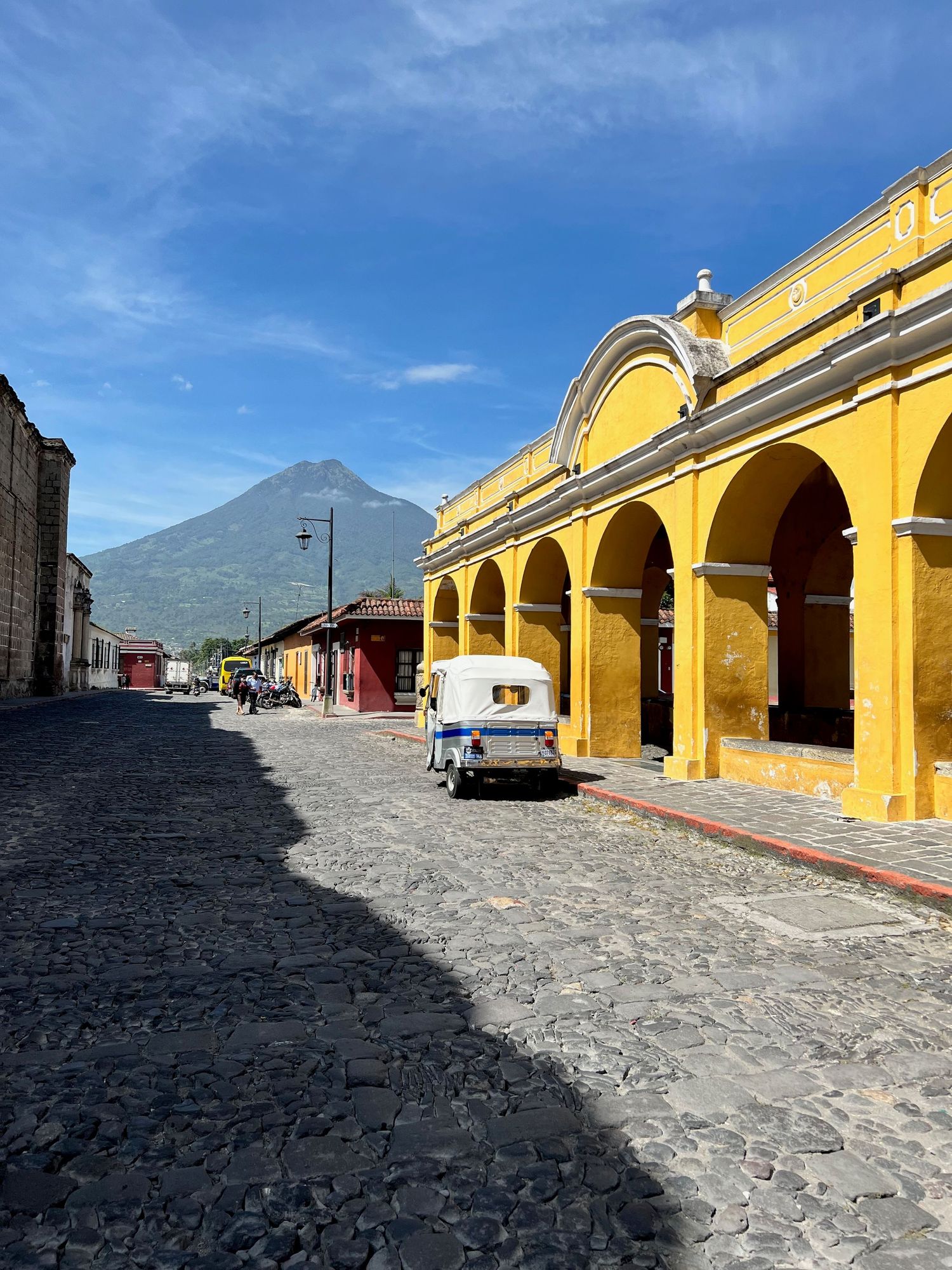 Cobblestone street in Antigua Guatemala with yellow colonial arcade building and Agua volcano in background.