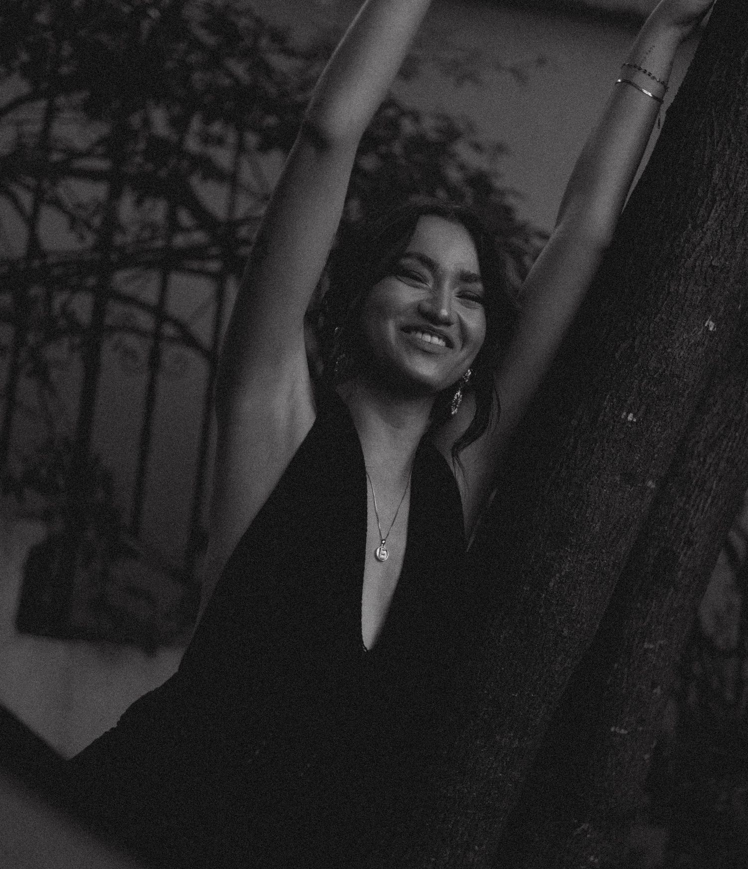 Black and white photo of a smiling woman in a black dress, arms raised, posing near a tree at night.