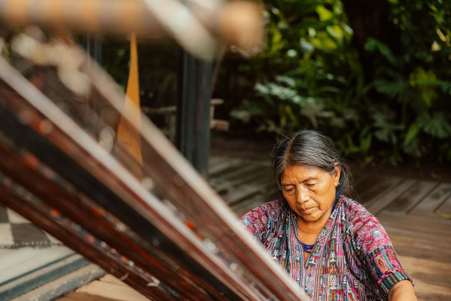 Indigenous woman weaving traditional textile on a backstrap loom outdoors in a lush green setting.