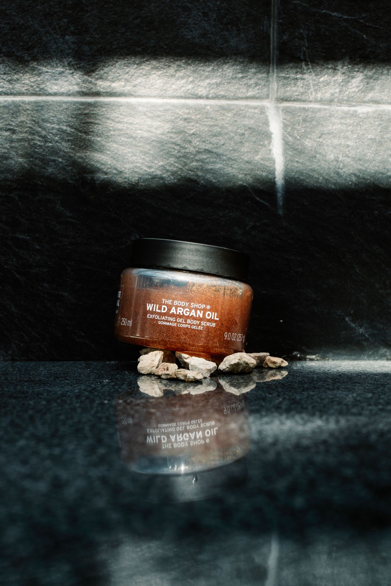 The Body Shop Wild Argan Oil exfoliating body scrub jar displayed on dark surface with scattered shells.