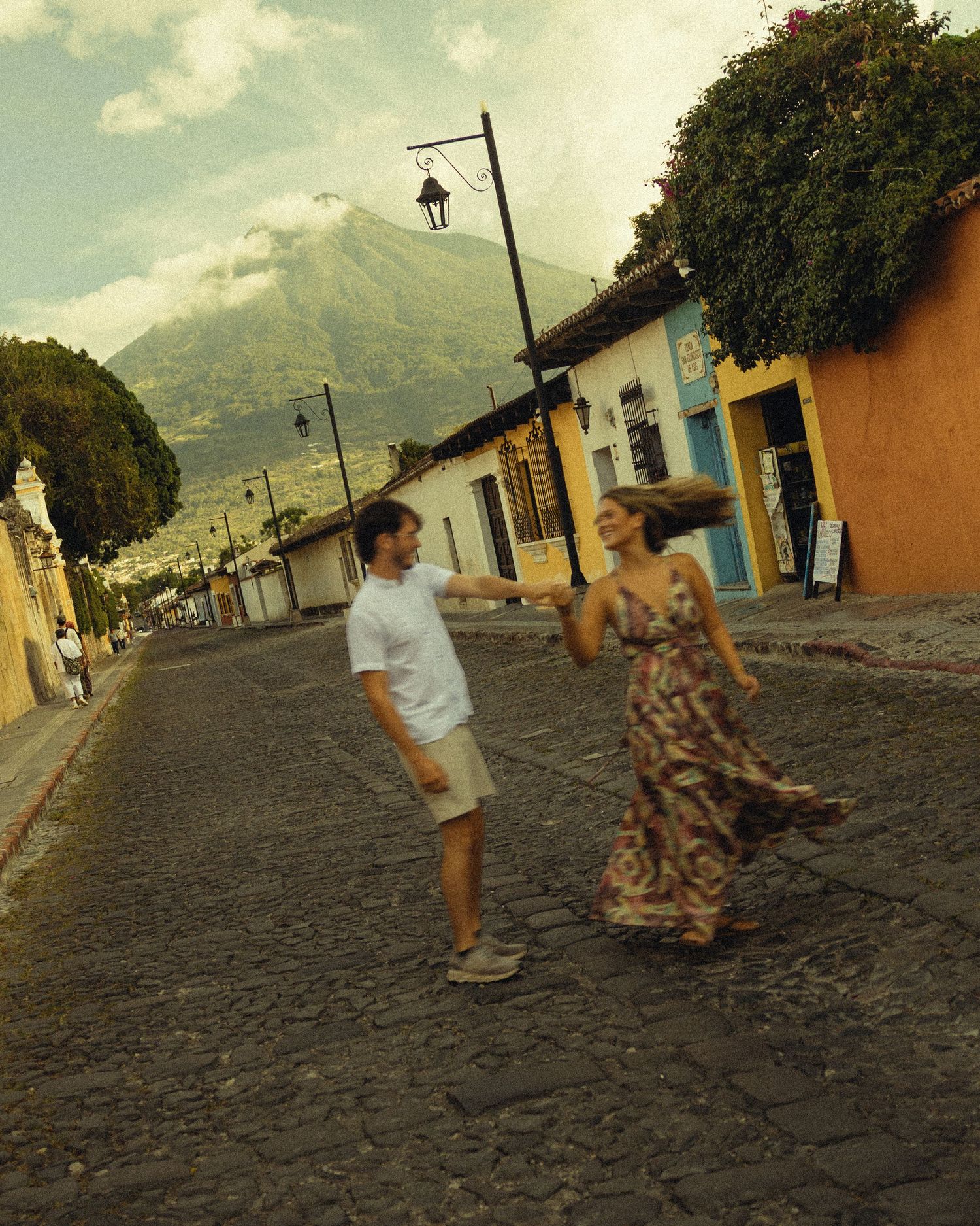 Couple dancing on cobblestone street in Antigua Guatemala with colorful buildings and volcano backdrop.