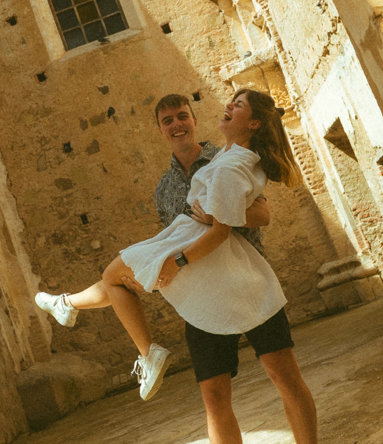 A happy couple posing together at an ancient stone landmark, man lifting woman in white dress outdoors.