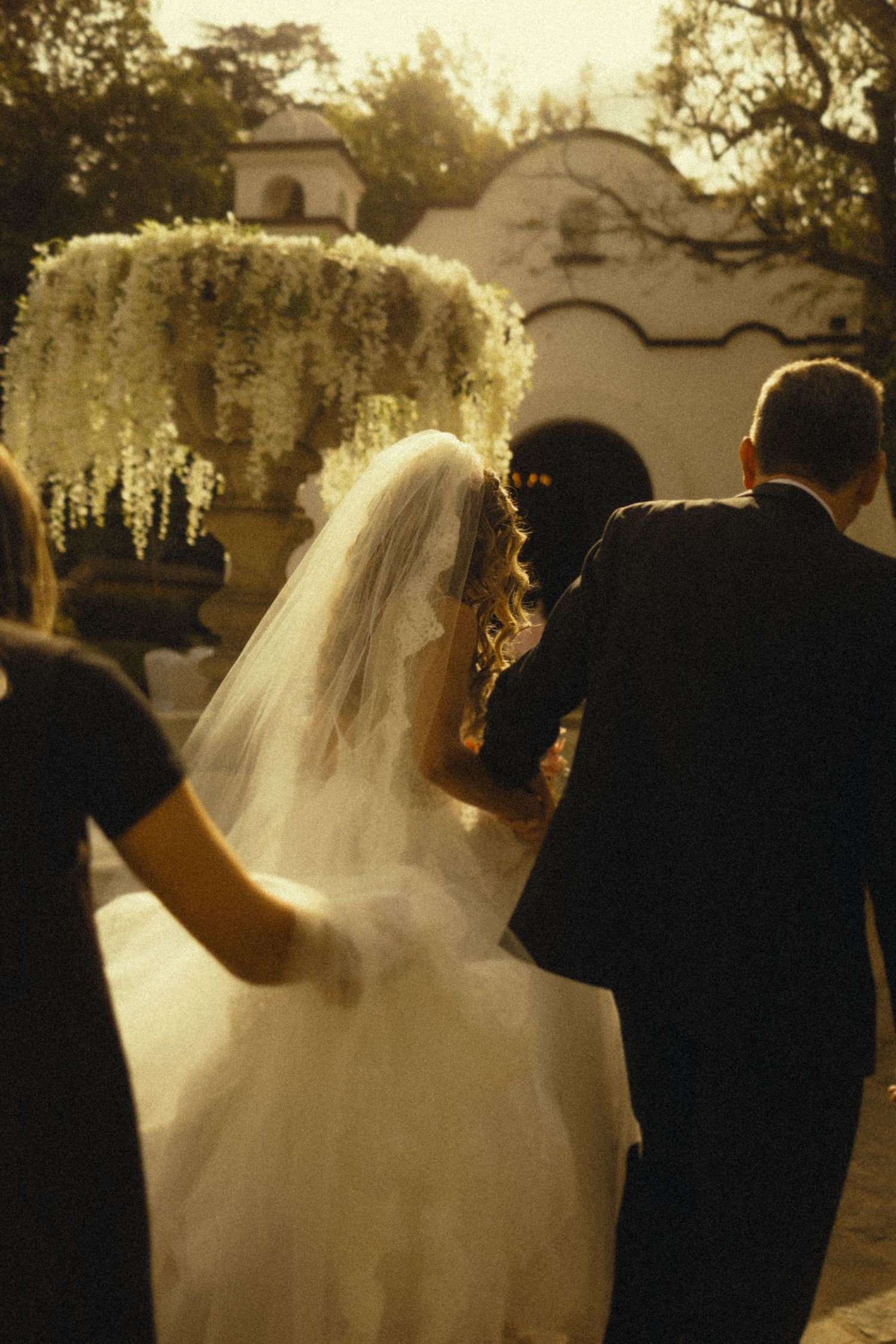 Bride in white gown and veil walks with groom toward floral arch at outdoor wedding ceremony.