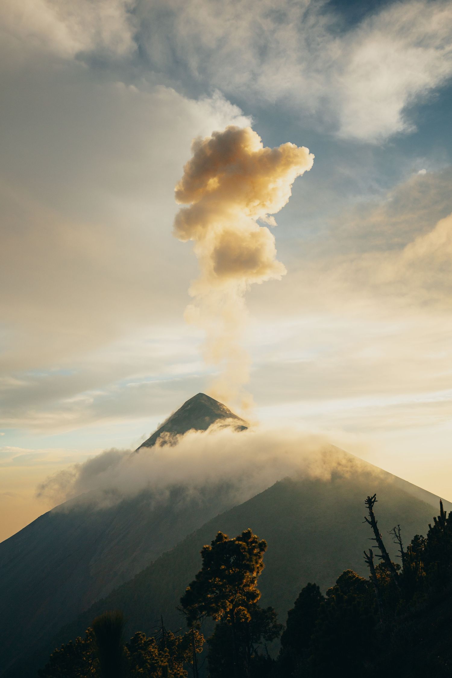 A volcanic mountain erupts with a billowing smoke plume rising into a golden cloudy sky at sunset.