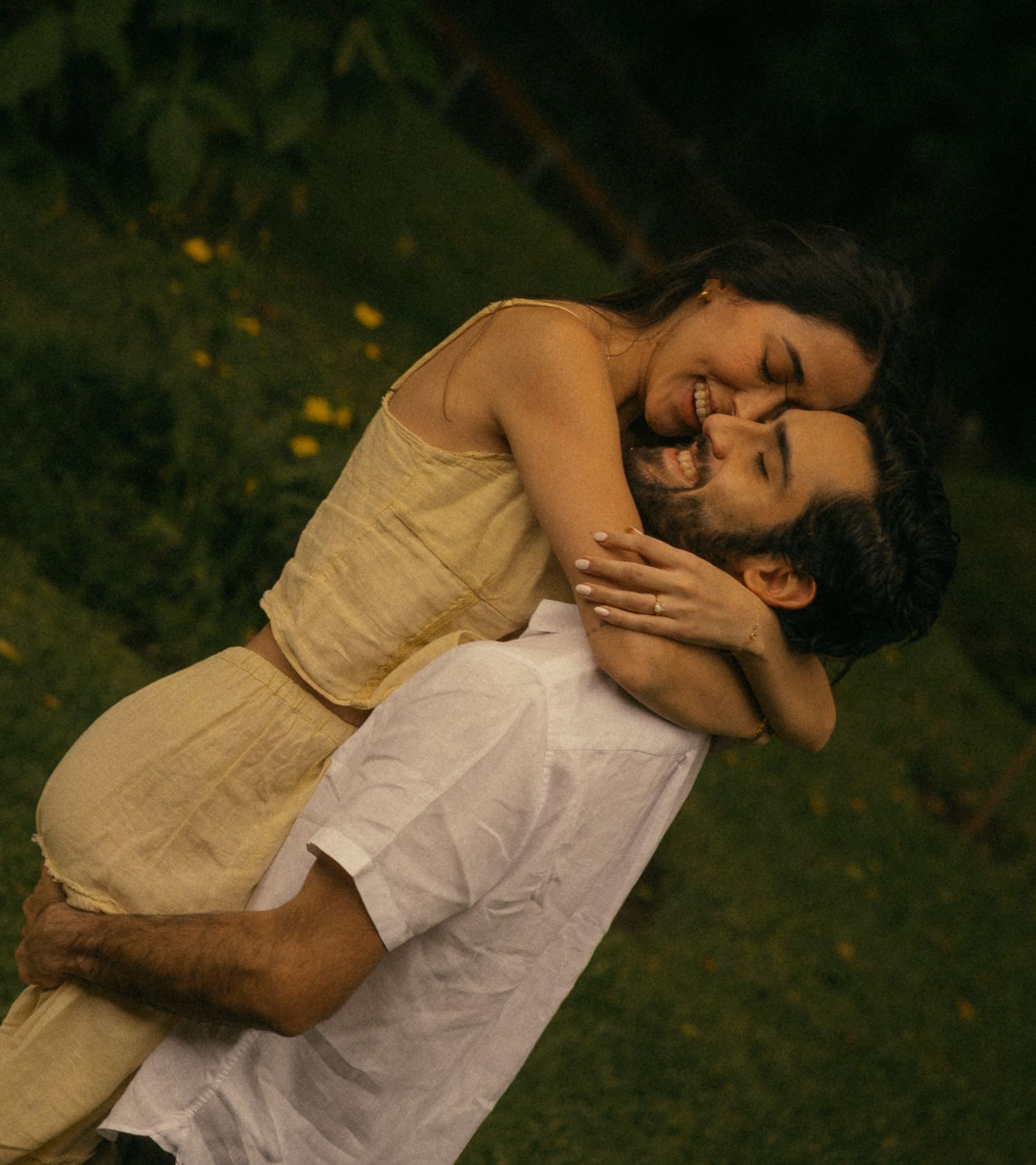 Joyful couple embracing outdoors, woman in yellow dress laughing as man lifts her in green garden.