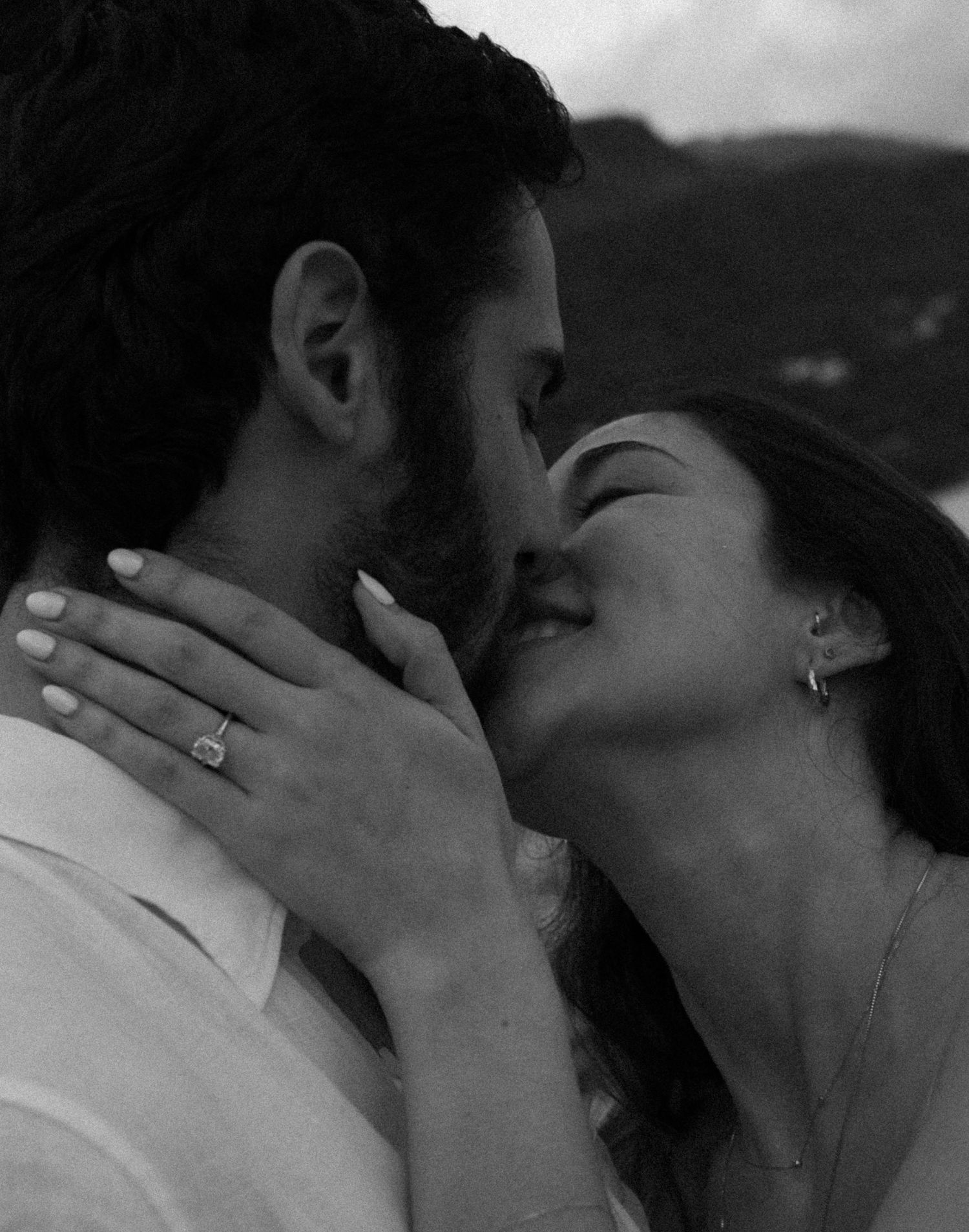 A couple shares a romantic kiss as the woman displays an elegant engagement ring in a black and white photo.