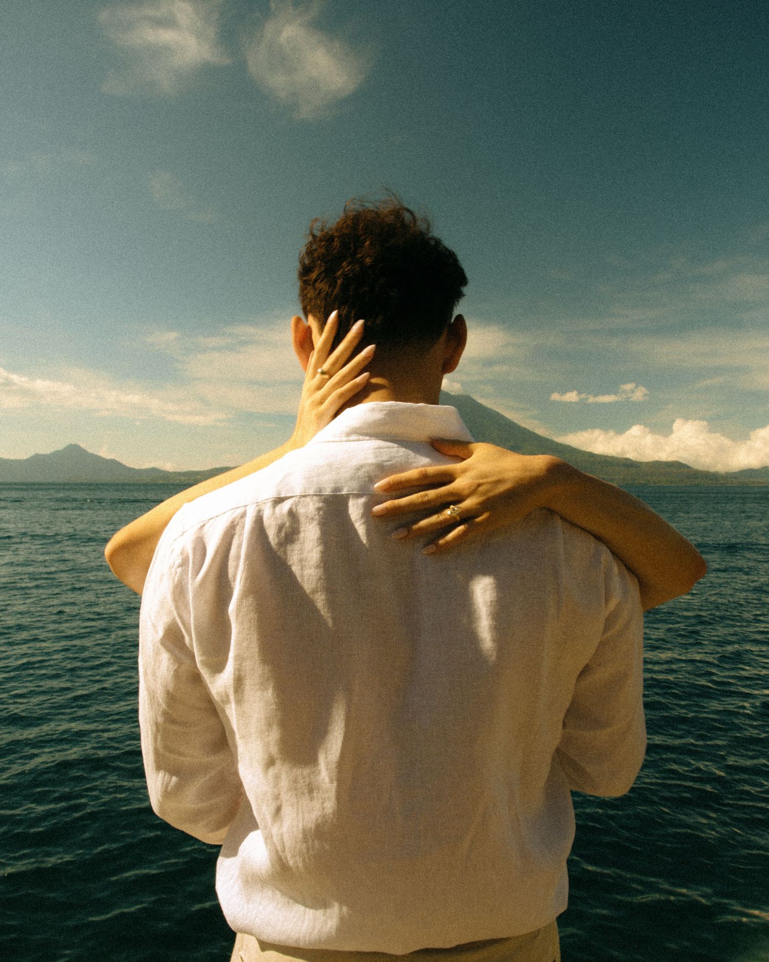 Two people embracing while gazing at a serene ocean view with mountains and cloudy sky in the background.