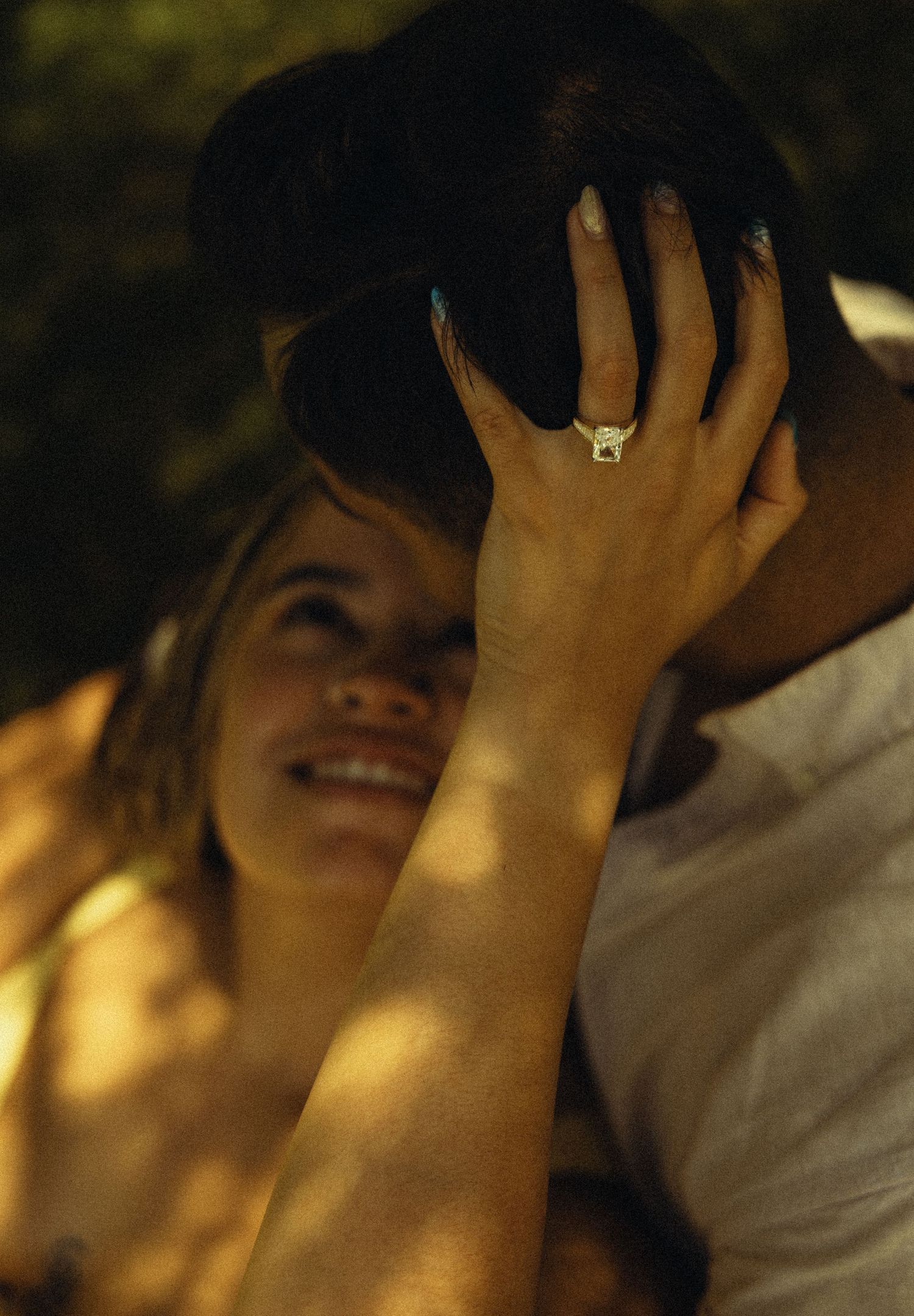 Smiling woman with engagement ring touching man's dark hair in a romantic outdoor setting.