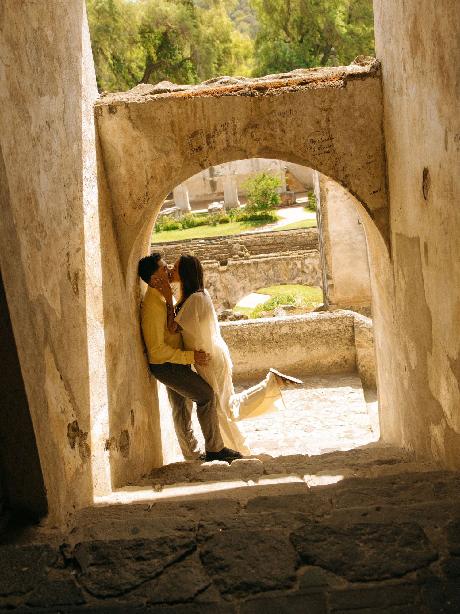 Couple embracing in a sunlit stone archway of a rustic historic building, with green gardens and old walls glowing in warm golden light.