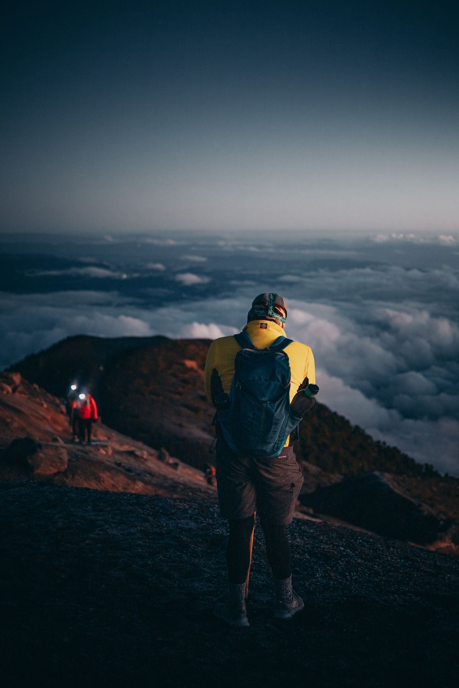 Hikers with headlamps descend a volcanic mountain ridge above a sea of clouds at dusk.
