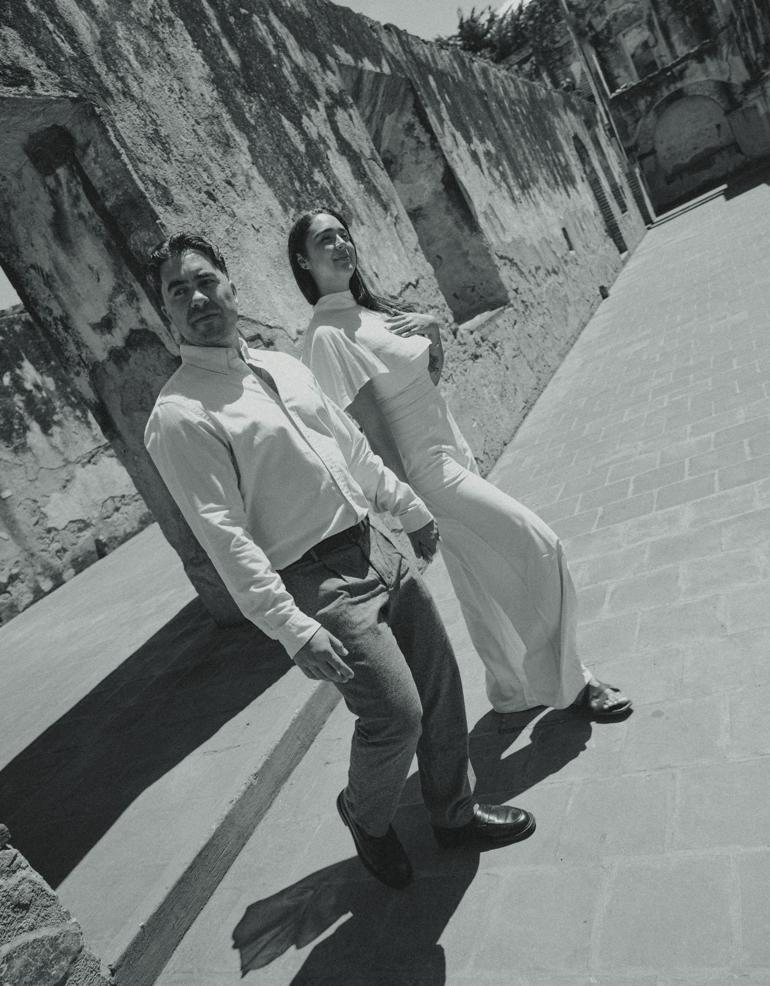Black and white photo of a couple in elegant attire posing on a sunlit cobblestone street beside a weathered stone wall.