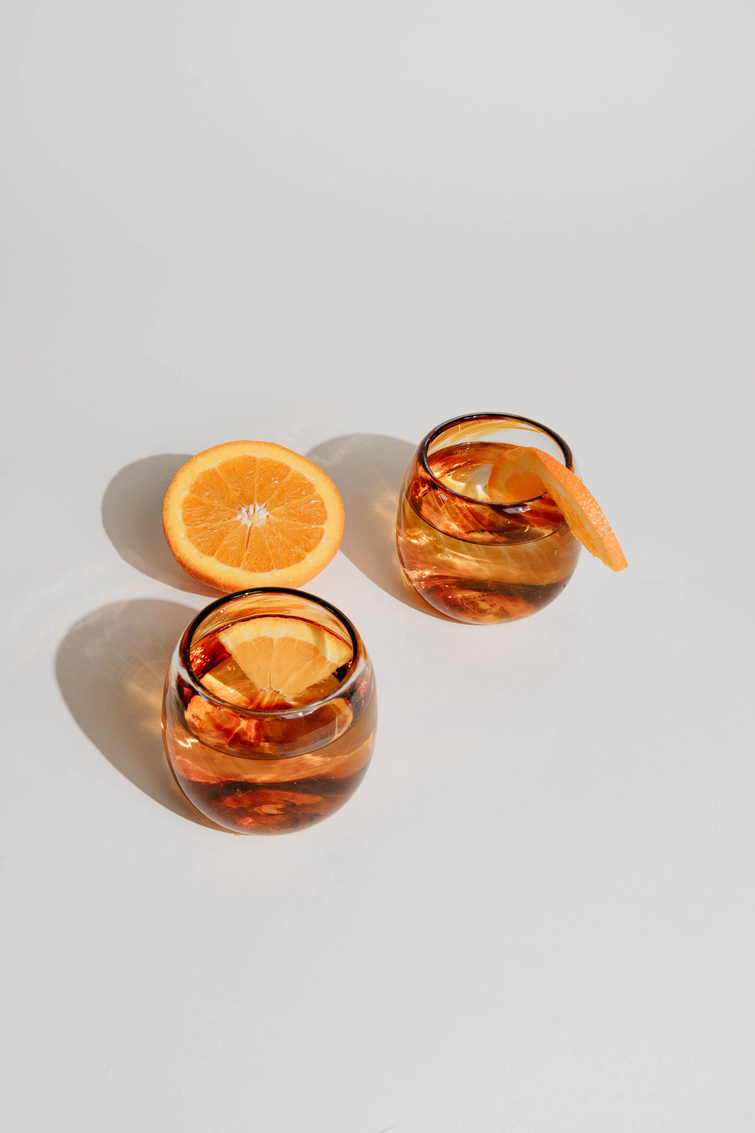 Two amber glass tumblers with orange slices and ice on white background with halved orange.