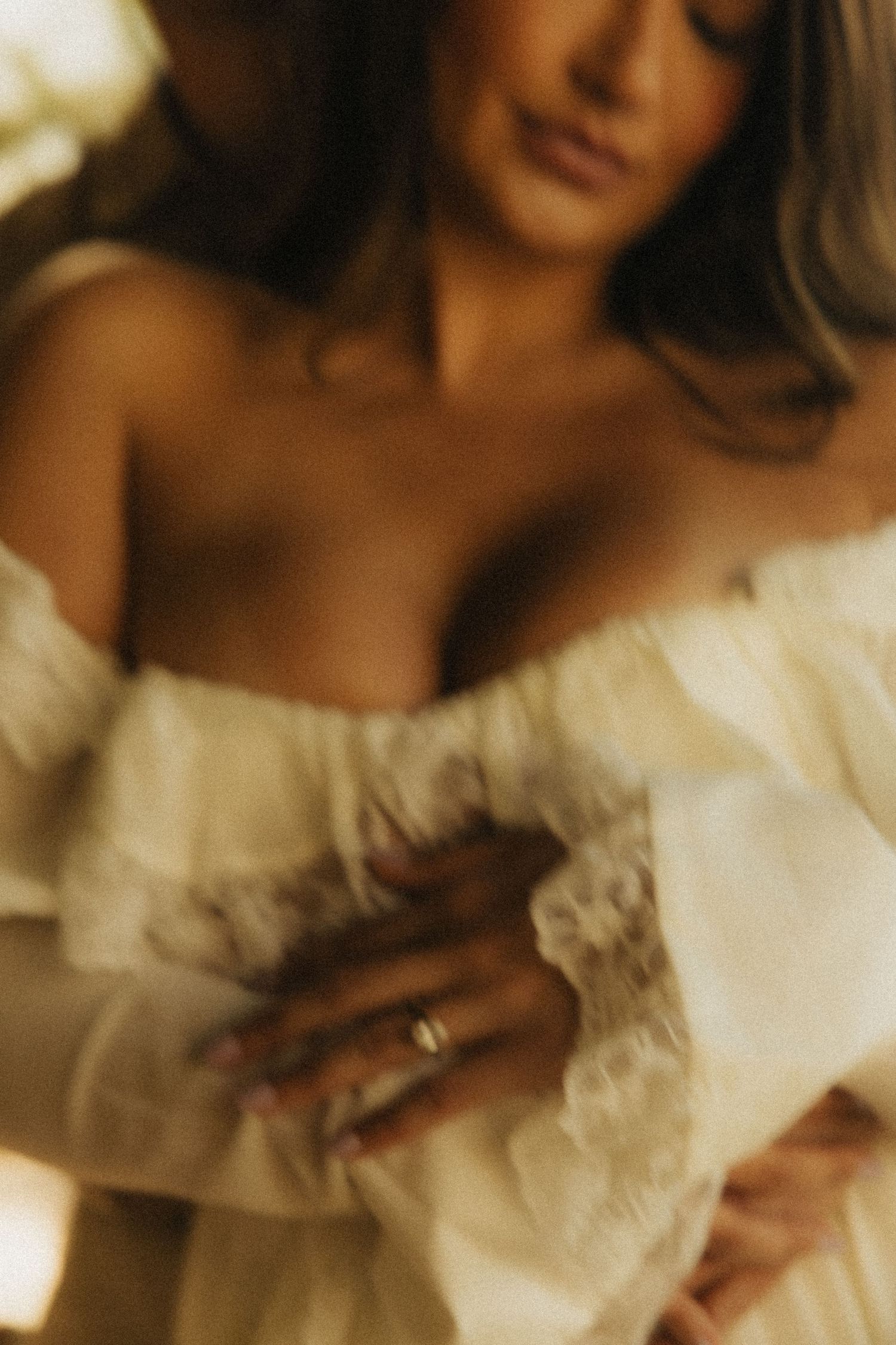Soft-focus portrait of a woman in a white off-shoulder lace gown, hands crossed over her chest.