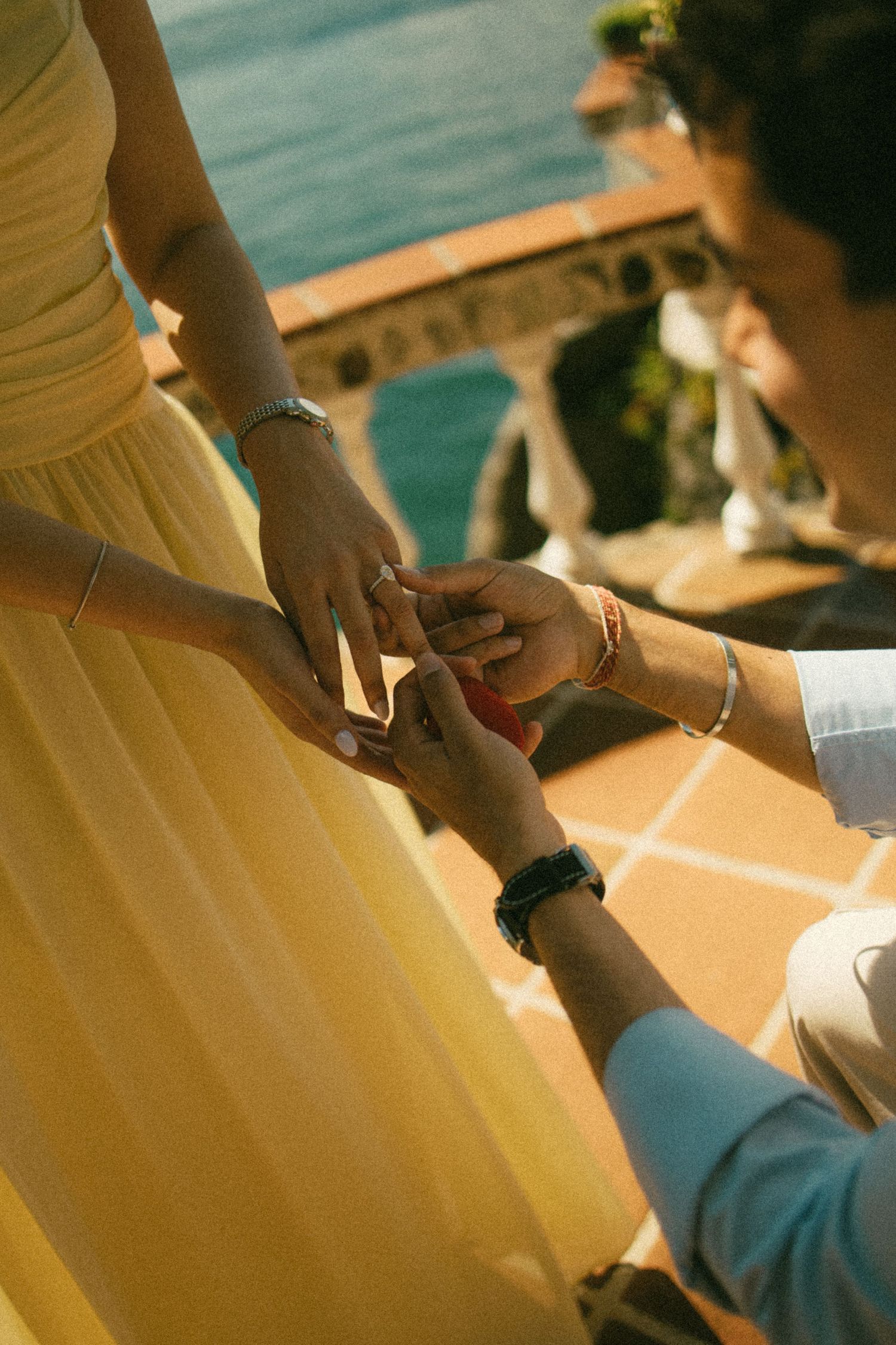A man kneels and slides an engagement ring onto a woman's finger on a sunny terrace overlooking the ocean.