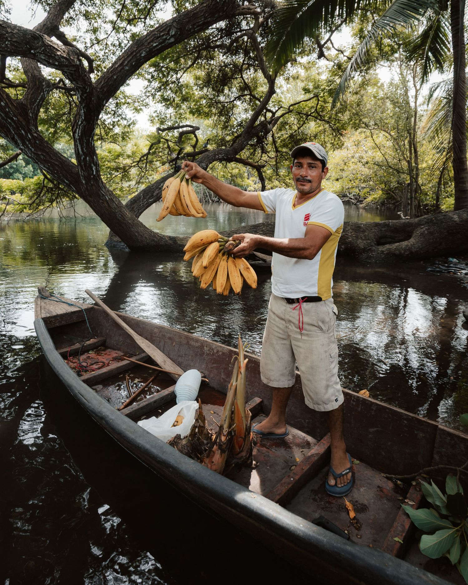 Man standing in a wooden canoe on a river holding fresh yellow bananas, surrounded by lush tropical trees.