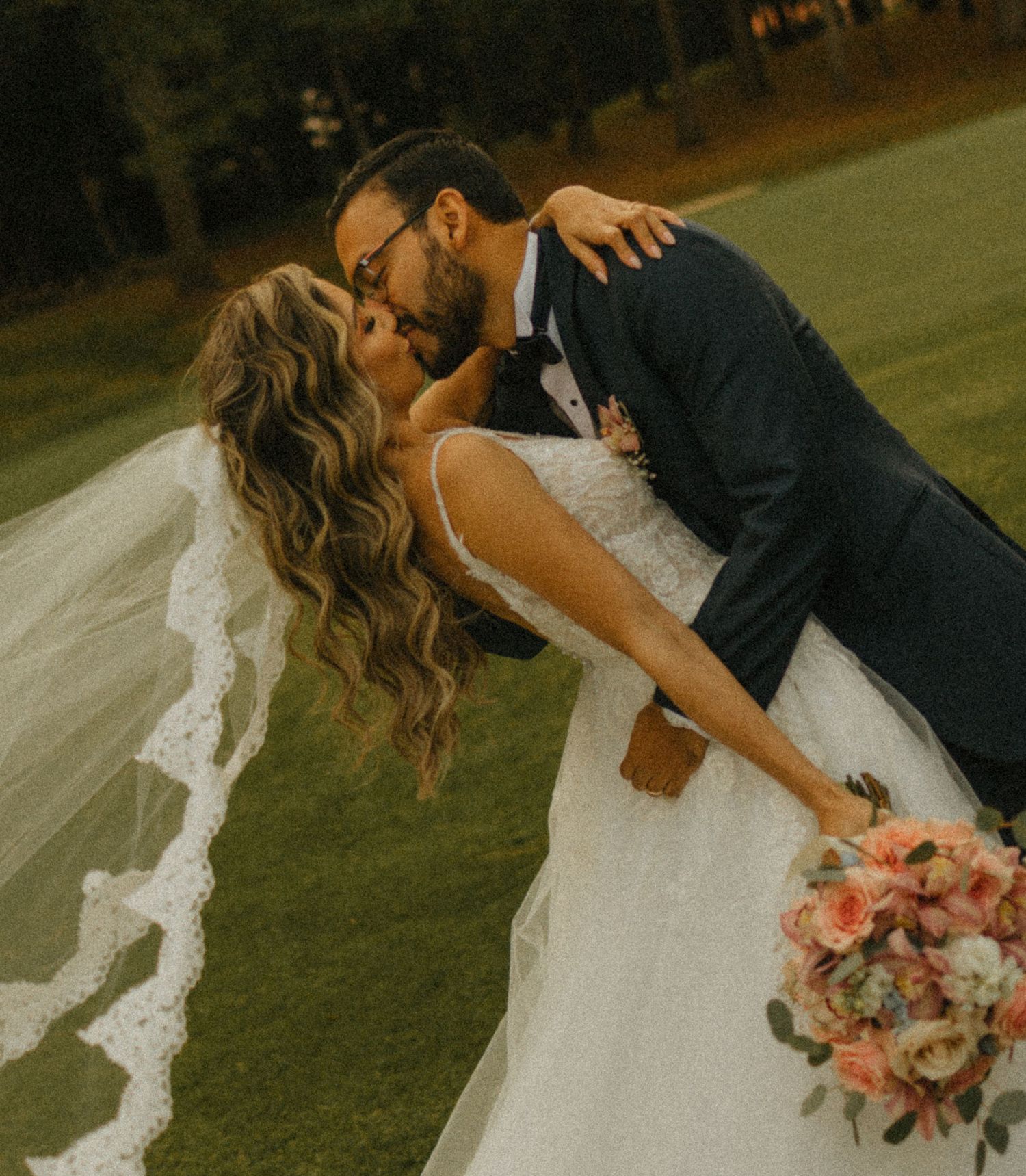 Bride and groom share a romantic dip kiss outdoors, bride holding pink bouquet wearing white gown with lace veil.
