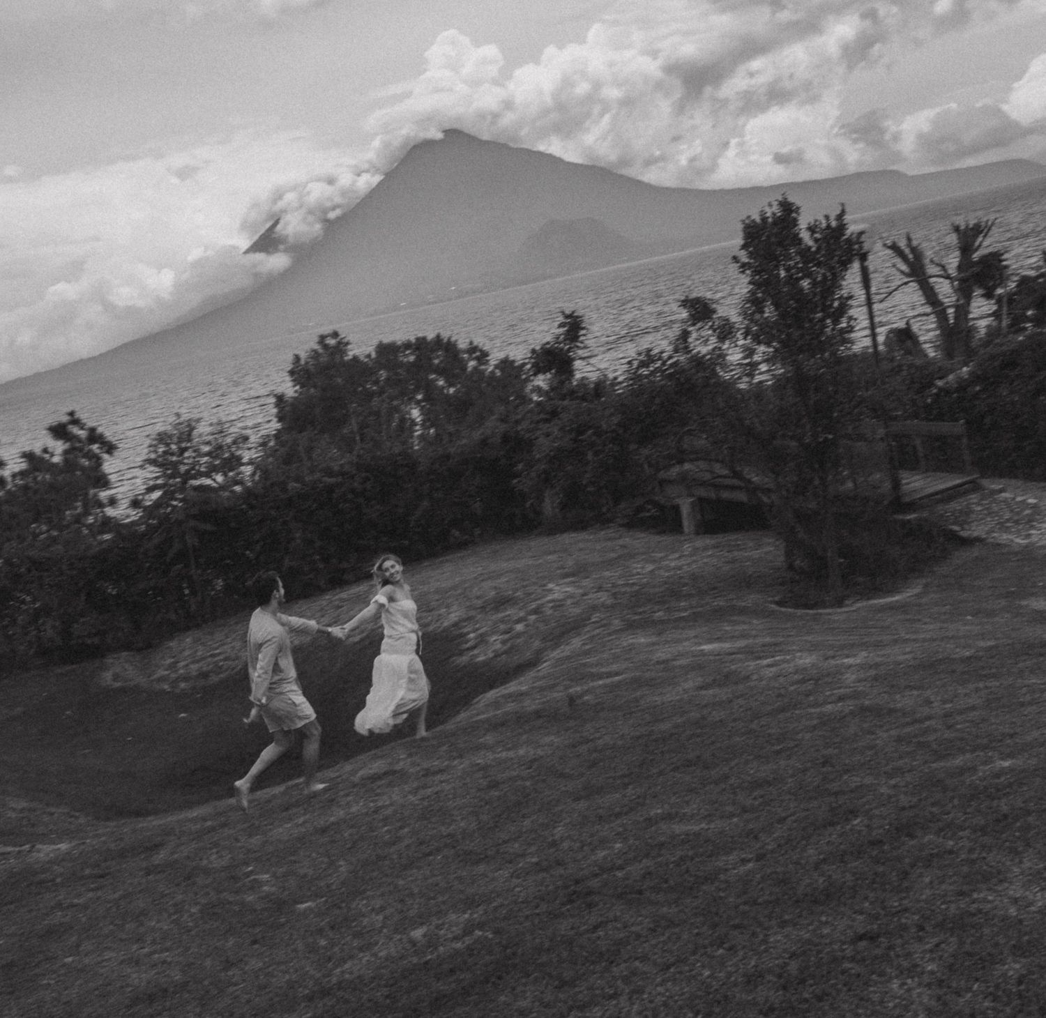 Two people dance holding hands on a grassy hillside with a volcano and dramatic clouds in a black and white photo.