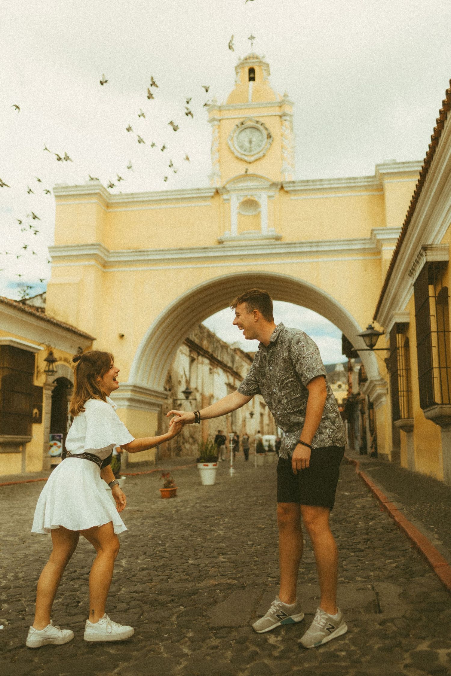 Couple holding hands on cobblestone street in Antigua Guatemala with iconic Santa Catalina Arch and clock tower behind them.