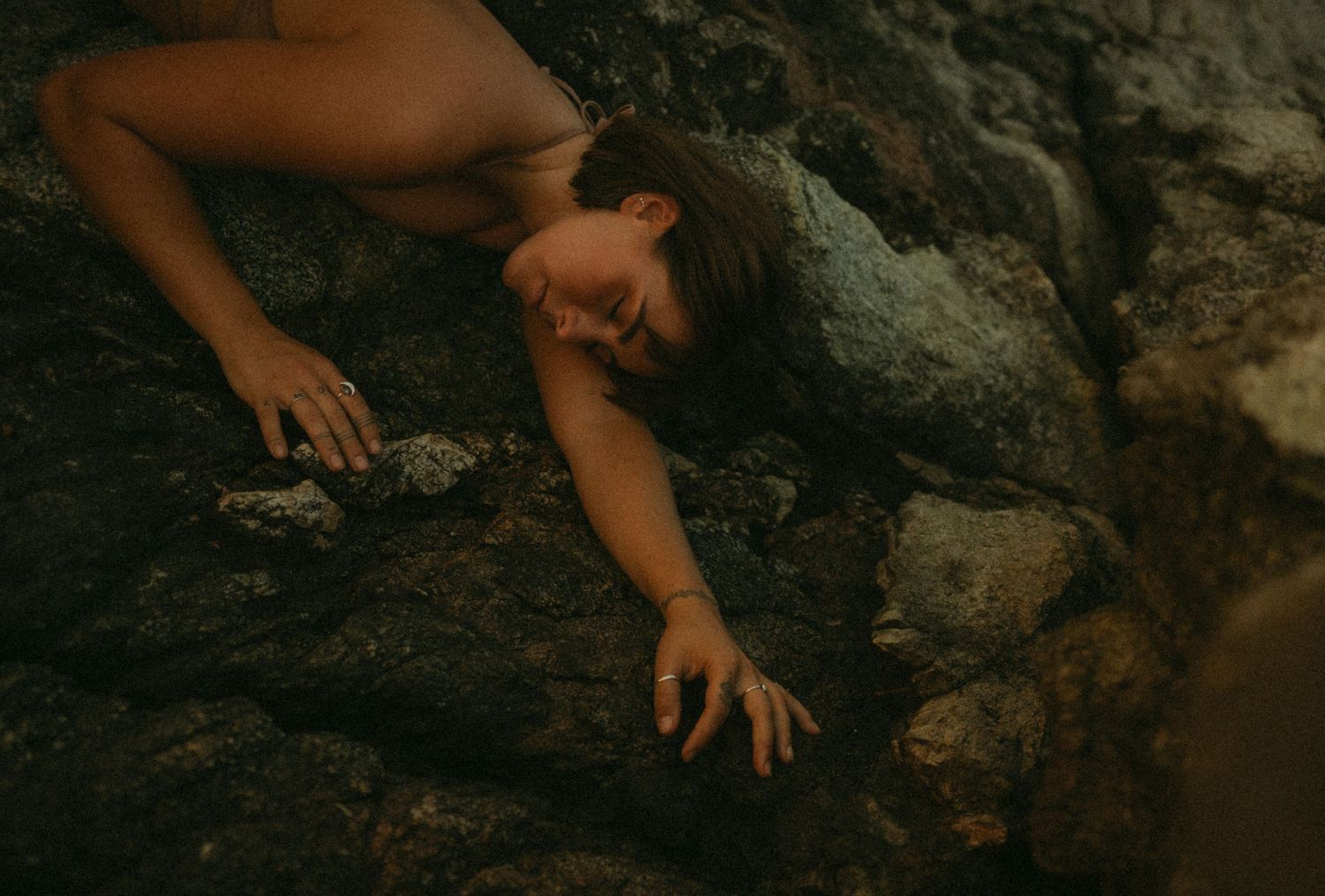 A woman lies dramatically across dark rocky terrain, reaching out with her hand in moody lighting.
