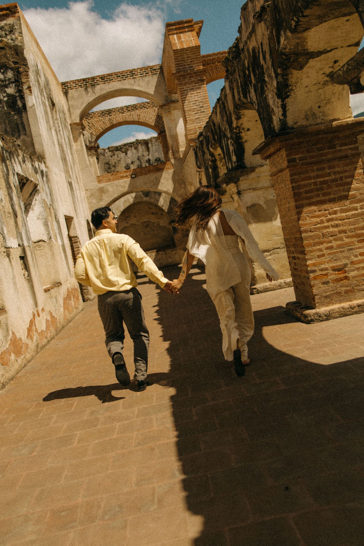 Couple holding hands walking through sunlit ancient brick archway ruins together.