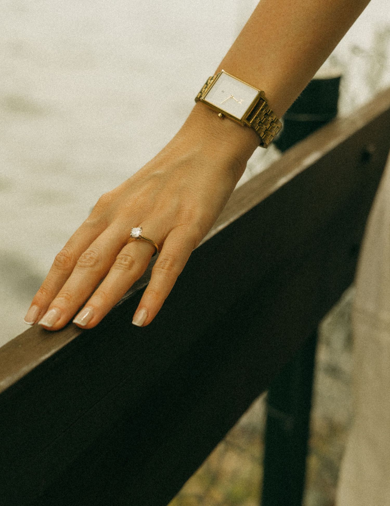 Close-up of a woman's hand wearing a gold watch and diamond solitaire ring, resting on a dark railing outdoors.