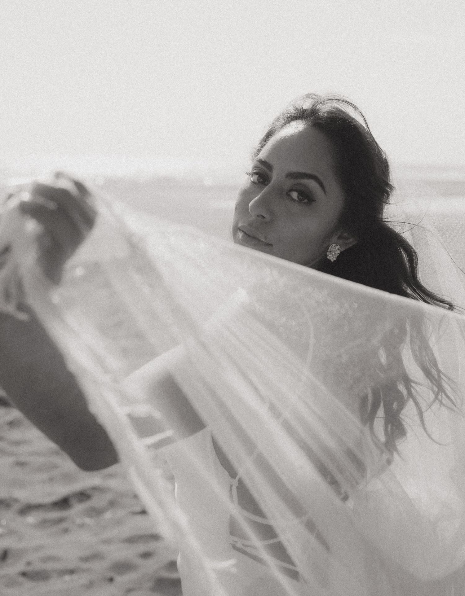 Black and white photo of a bride holding her veil on a beach, looking over her shoulder with elegant earrings.