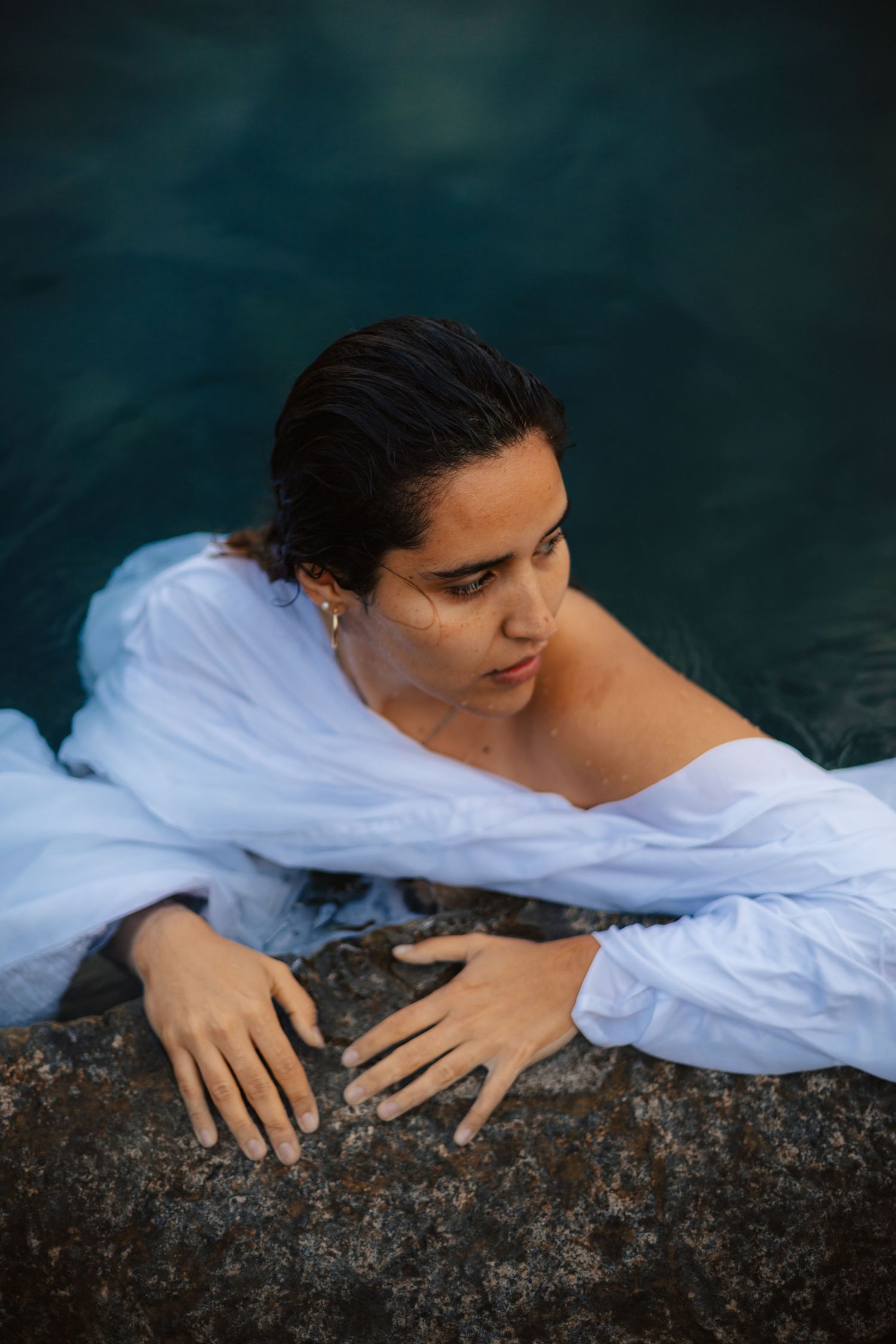 Woman in wet white shirt resting on rocky pool edge in turquoise water, gazing thoughtfully to the side.