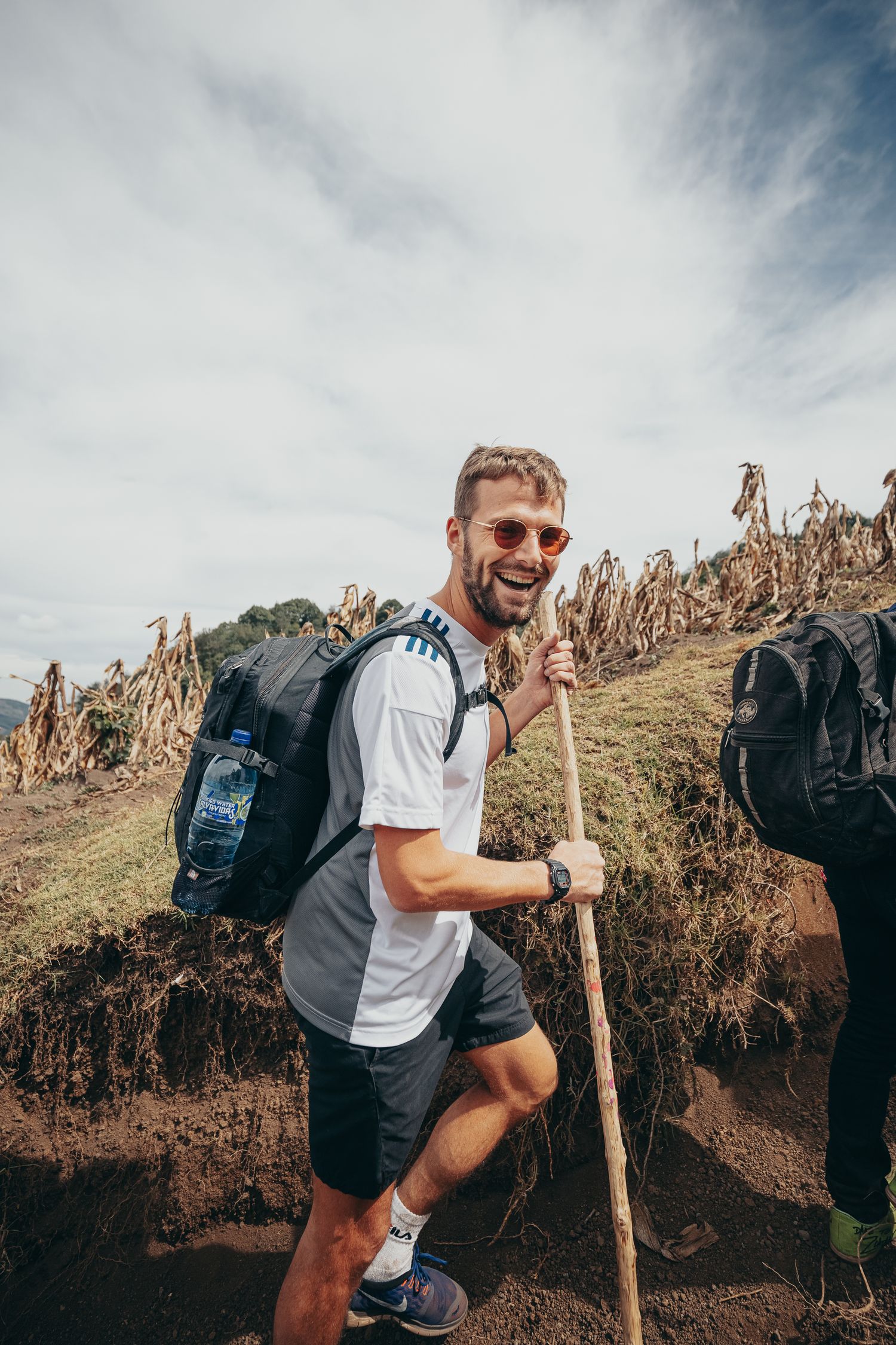 Smiling male hiker with backpack and walking stick trekking on a mountain trail under a cloudy sky.