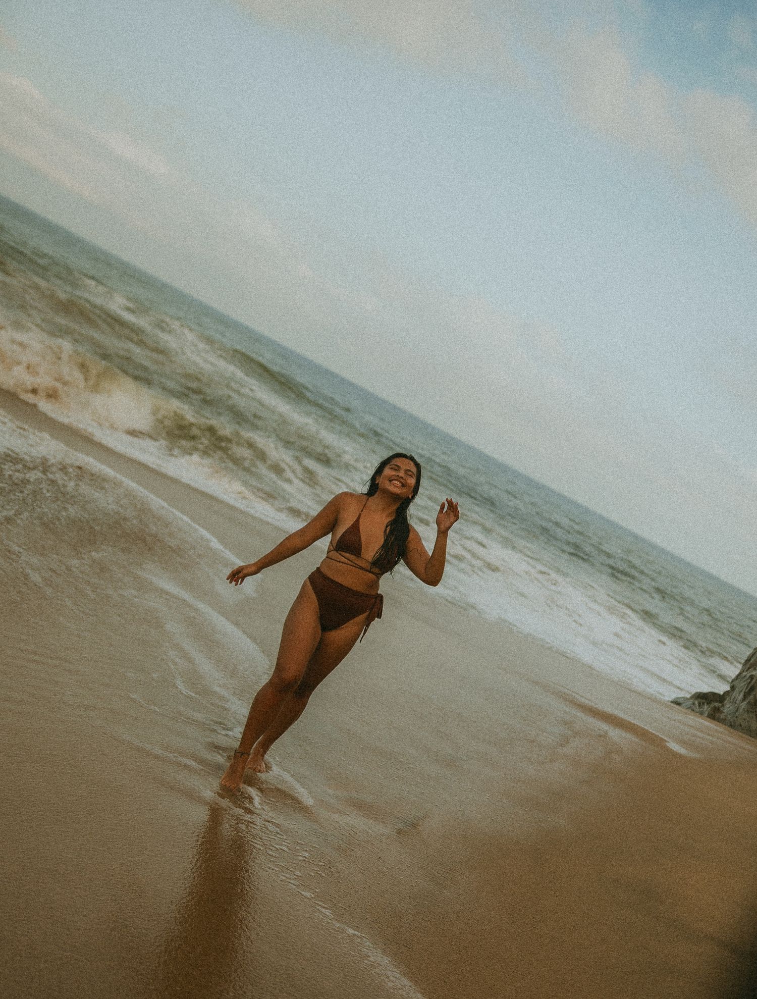 Woman in brown bikini enjoying ocean waves on a sandy beach under a cloudy sky.