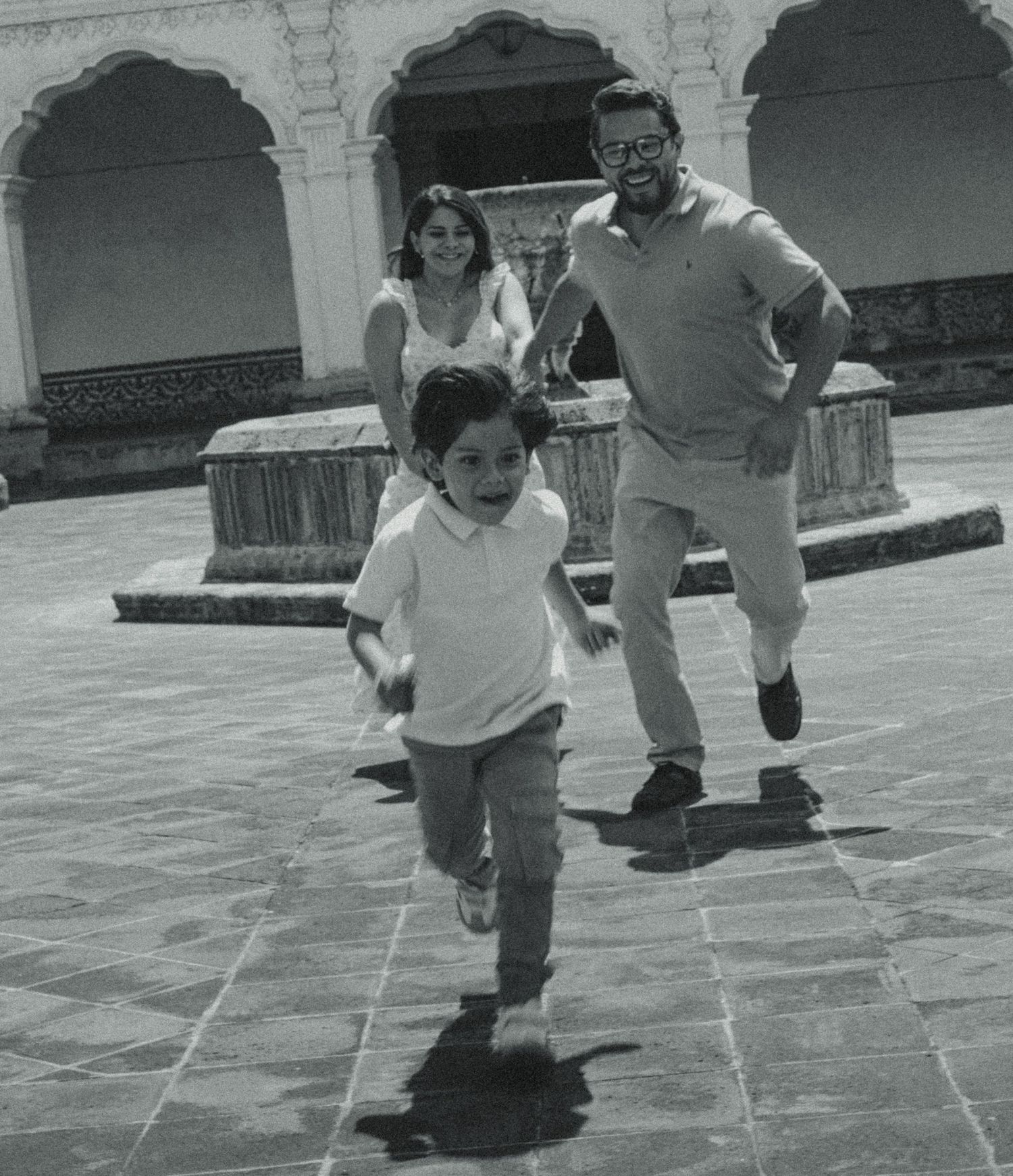 A joyful child runs ahead of a smiling couple in a sunlit courtyard with arched architecture.