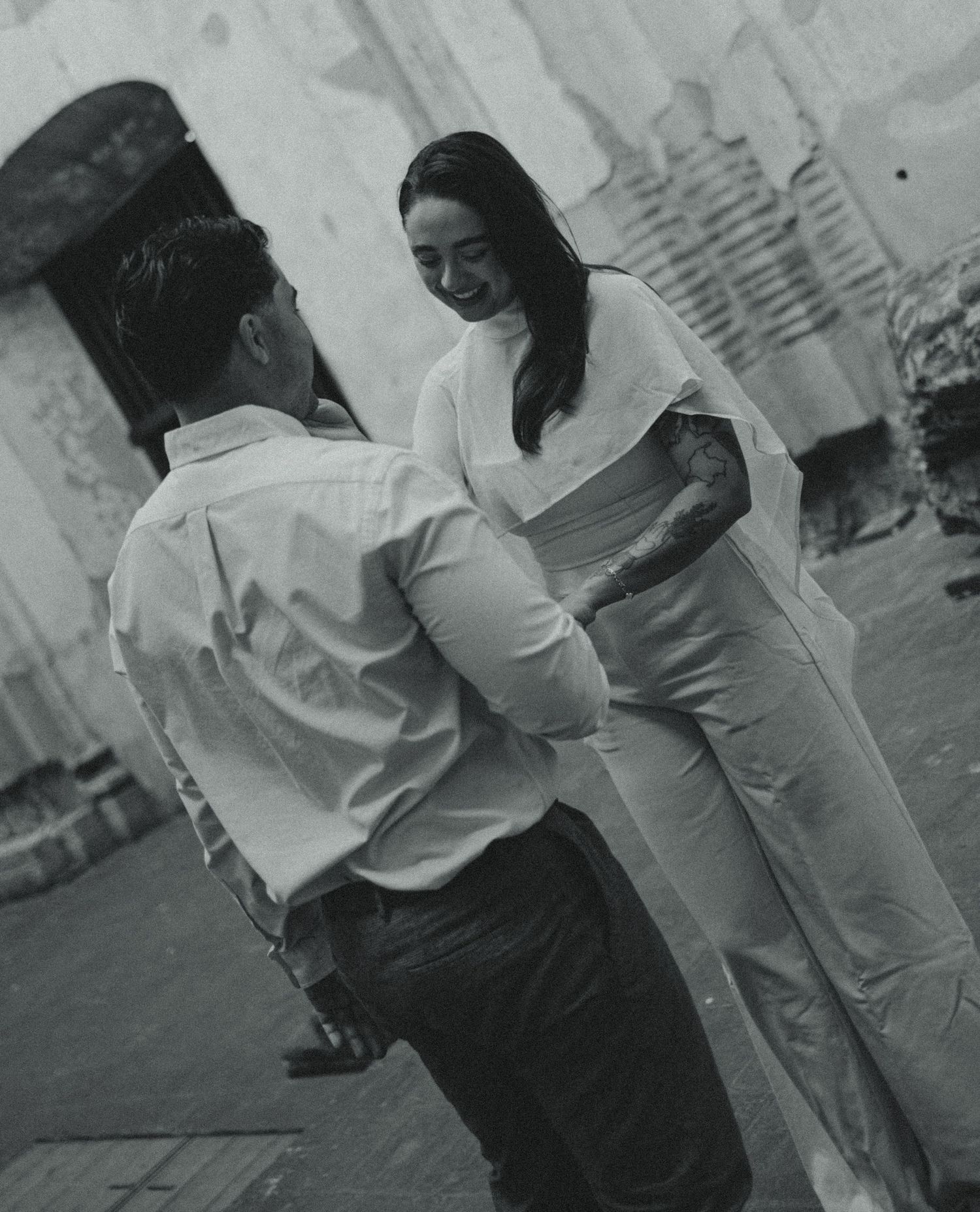 Black and white photo of a smiling woman in a white jumpsuit facing a man in a button-up shirt outside a weathered stone building.