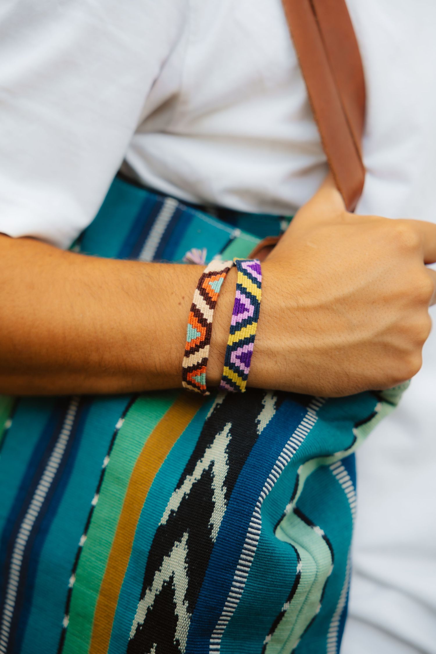 Close-up of woven geometric friendship bracelets on wrist holding a teal striped bag with leather strap.