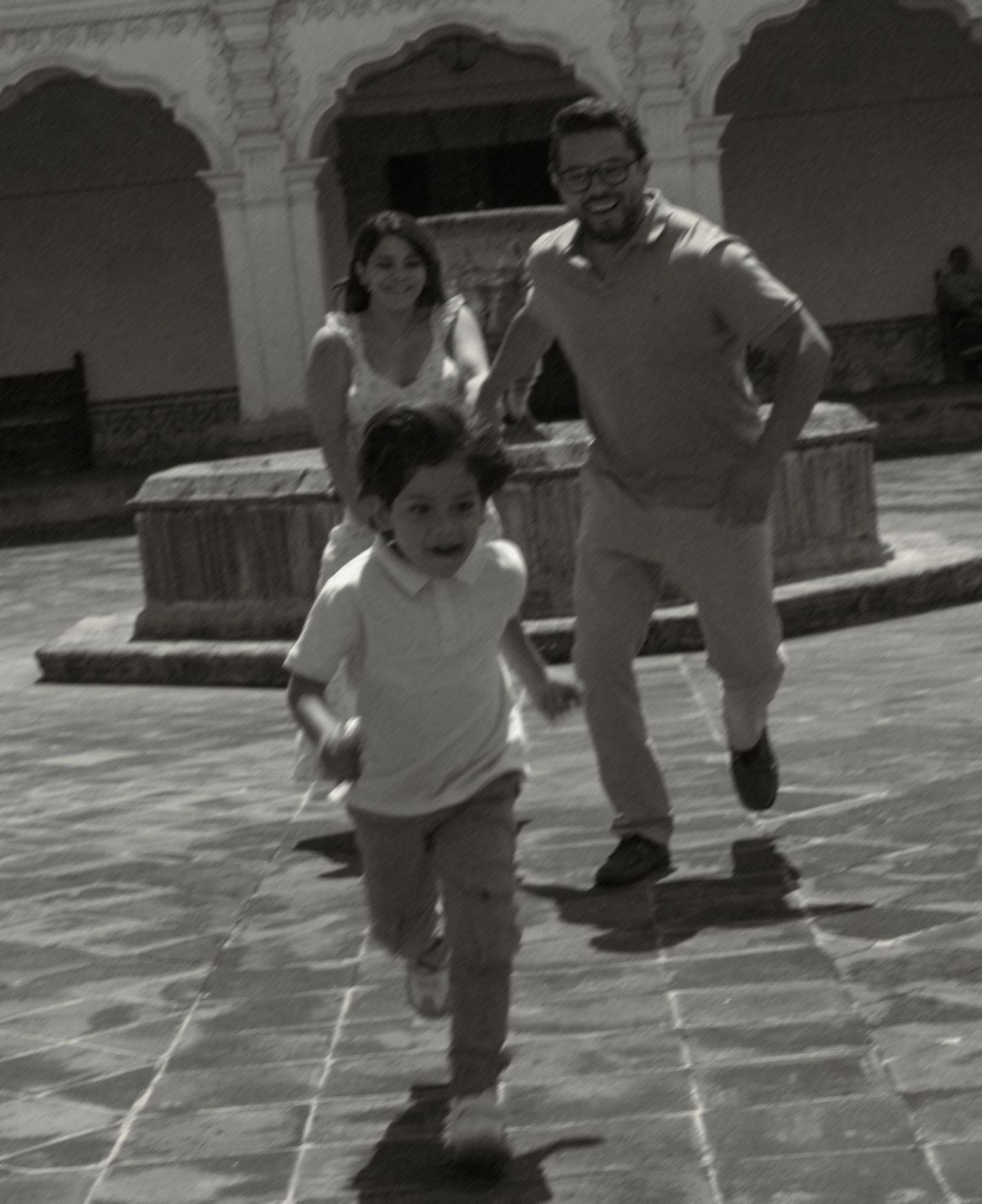 Black and white photo of a joyful family running in a courtyard, with a child leading and parents smiling behind.