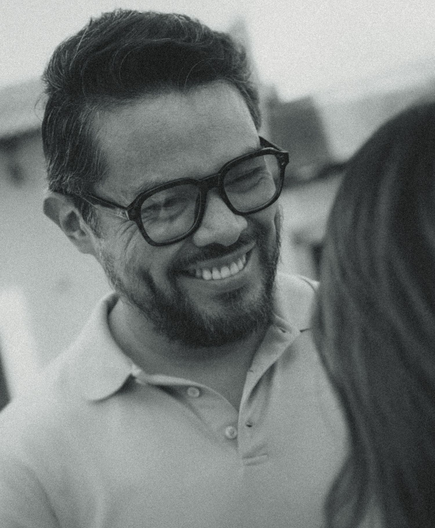 Smiling bearded man with black glasses enjoying a conversation outdoors in a candid black and white photo.