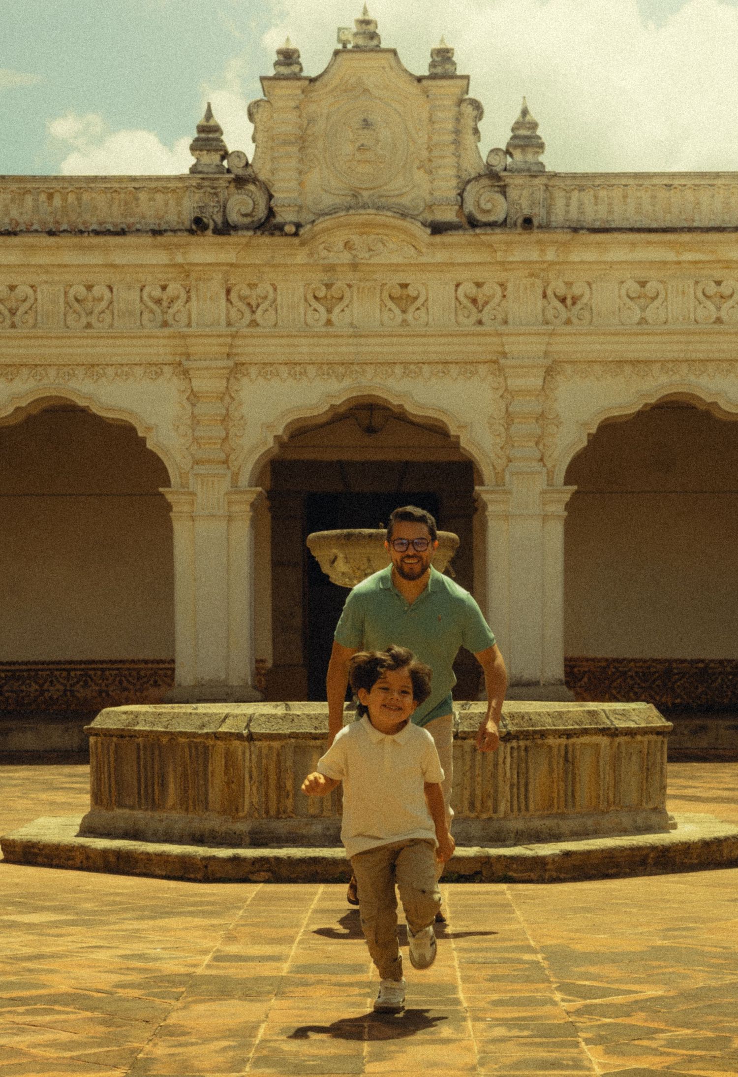 A child and man run through a courtyard of a historic colonial building with arched walkways and a fountain.