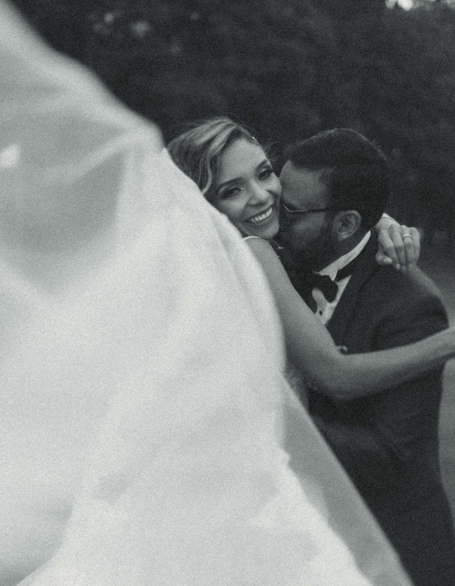 Black and white wedding photo of smiling bride and groom embracing outdoors, with flowing veil in foreground.