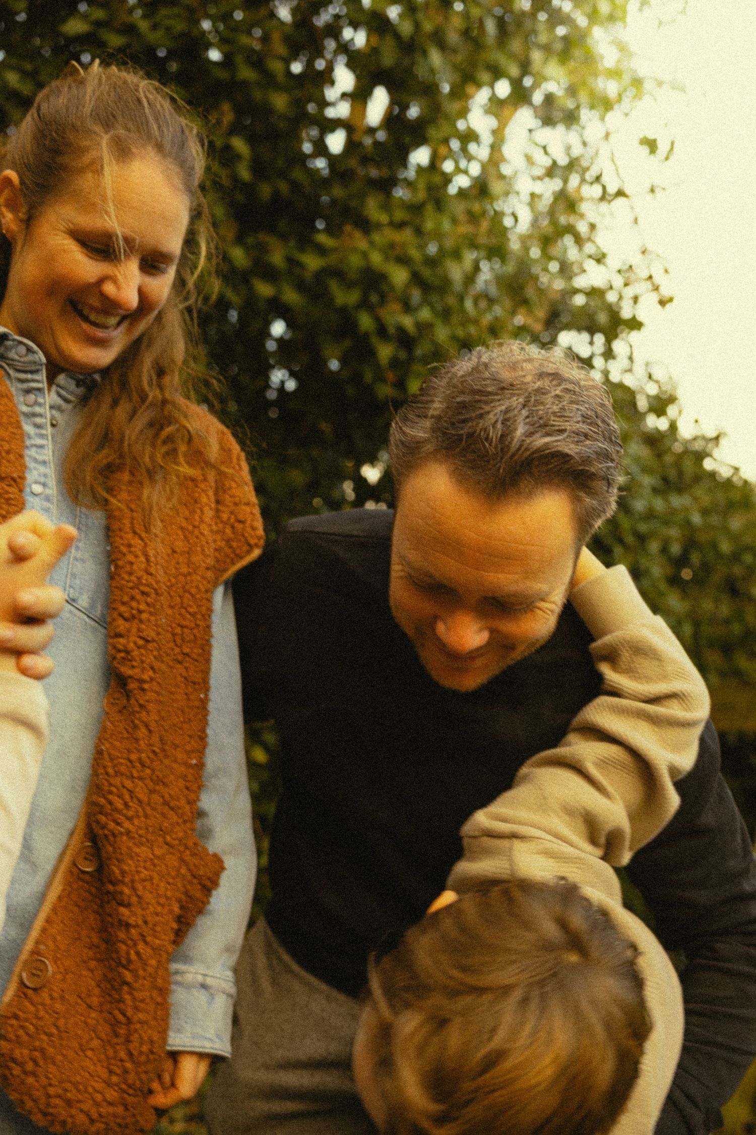 A smiling family with two parents and a child enjoying a playful outdoor moment surrounded by green trees.