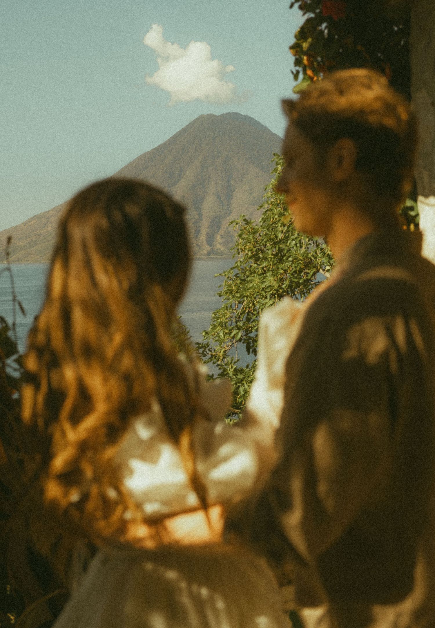 Couple gazing at a volcanic mountain over Lake Atitlan, Guatemala, surrounded by lush greenery on a sunny day.